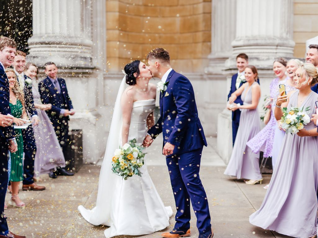 A bride and groom kiss whilst brightly coloured confetti floats in the air around them