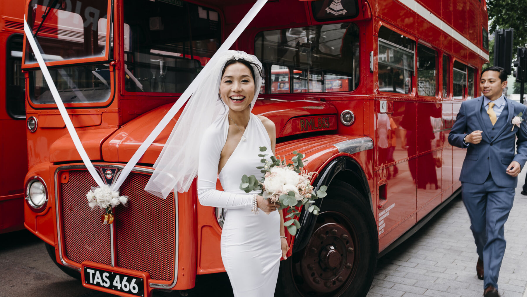 Chinese bride in one shoulder wedding dress standing next to red London bus. Sandra Reddin Photography: London wedding photographer