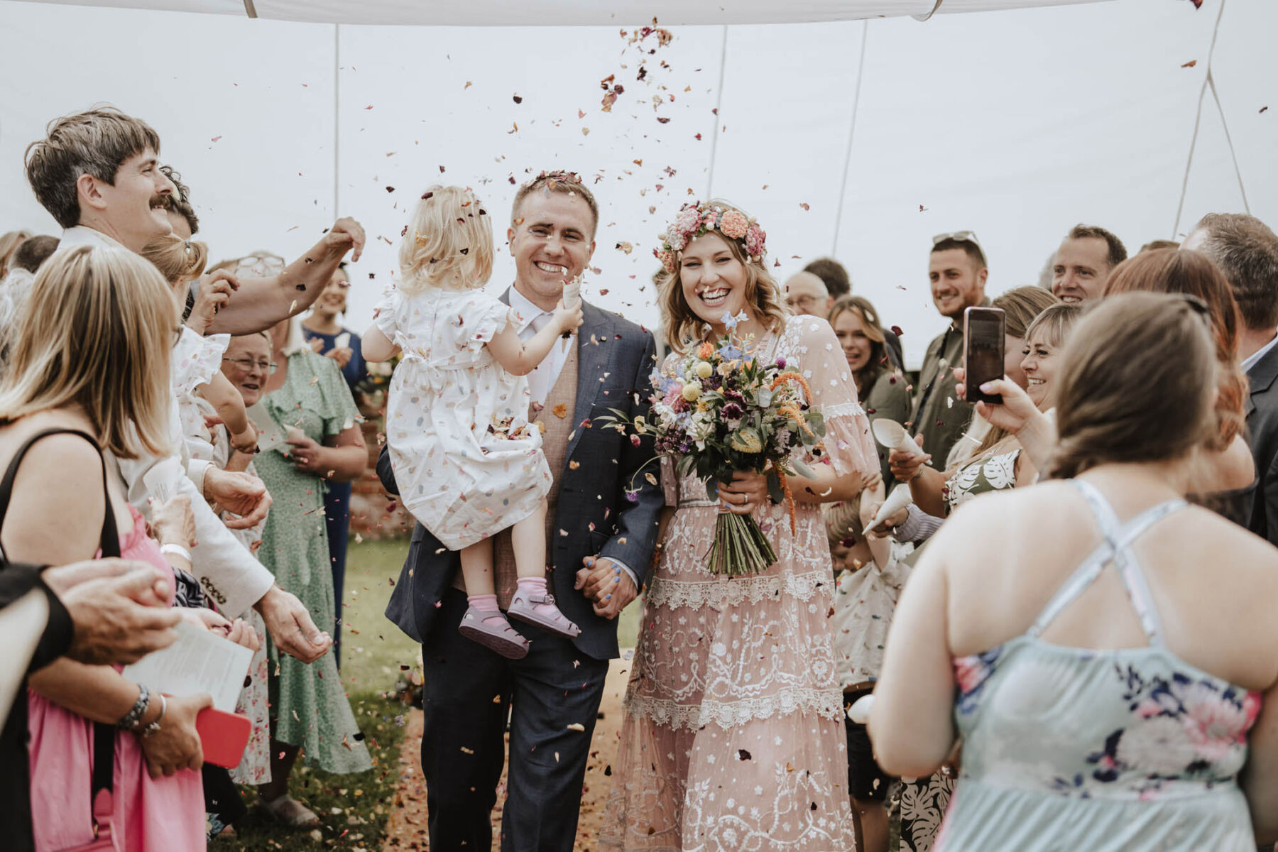 Bride in pale pink Needle & Thread wedding dress. Confetti shot. Groom carries little flower girl in his arms.