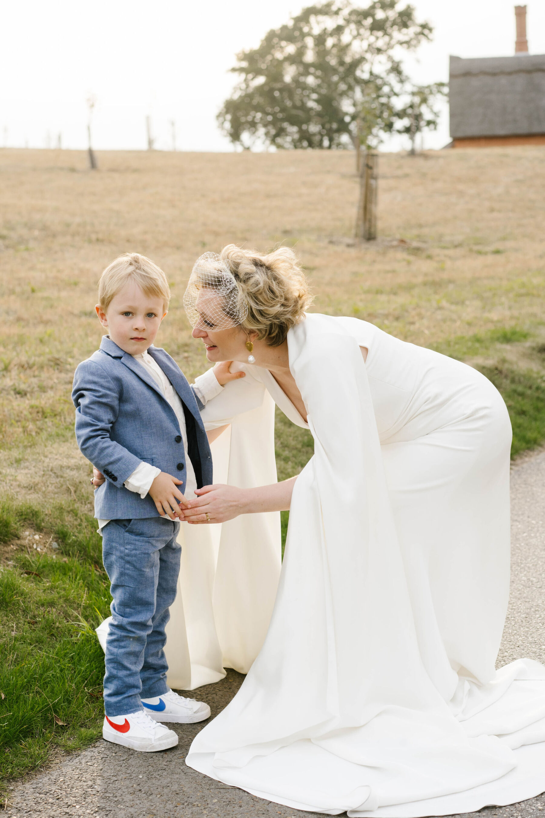 Andrea Hawkes bride, birdcage veil. Bride bending to comfort page boy in pale blue suit + Nike trainers.