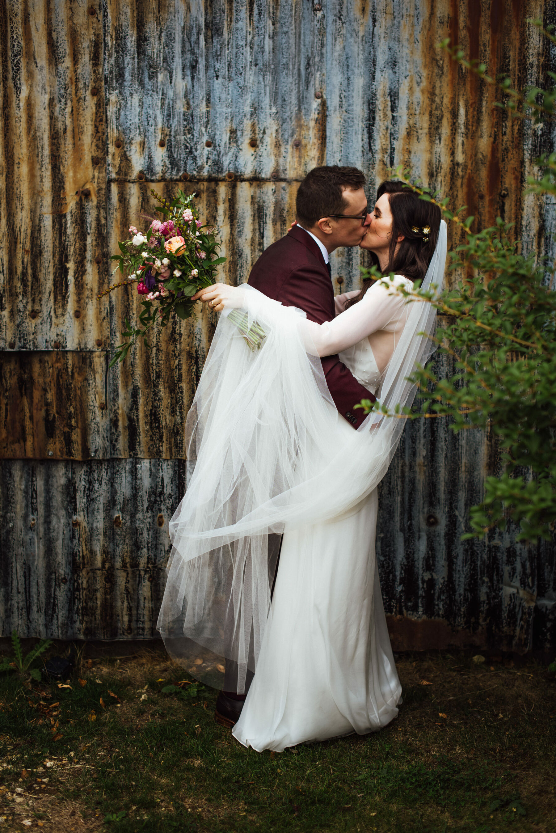 Andrea Hawkes bride kissing her groom in a burgundy suit.
