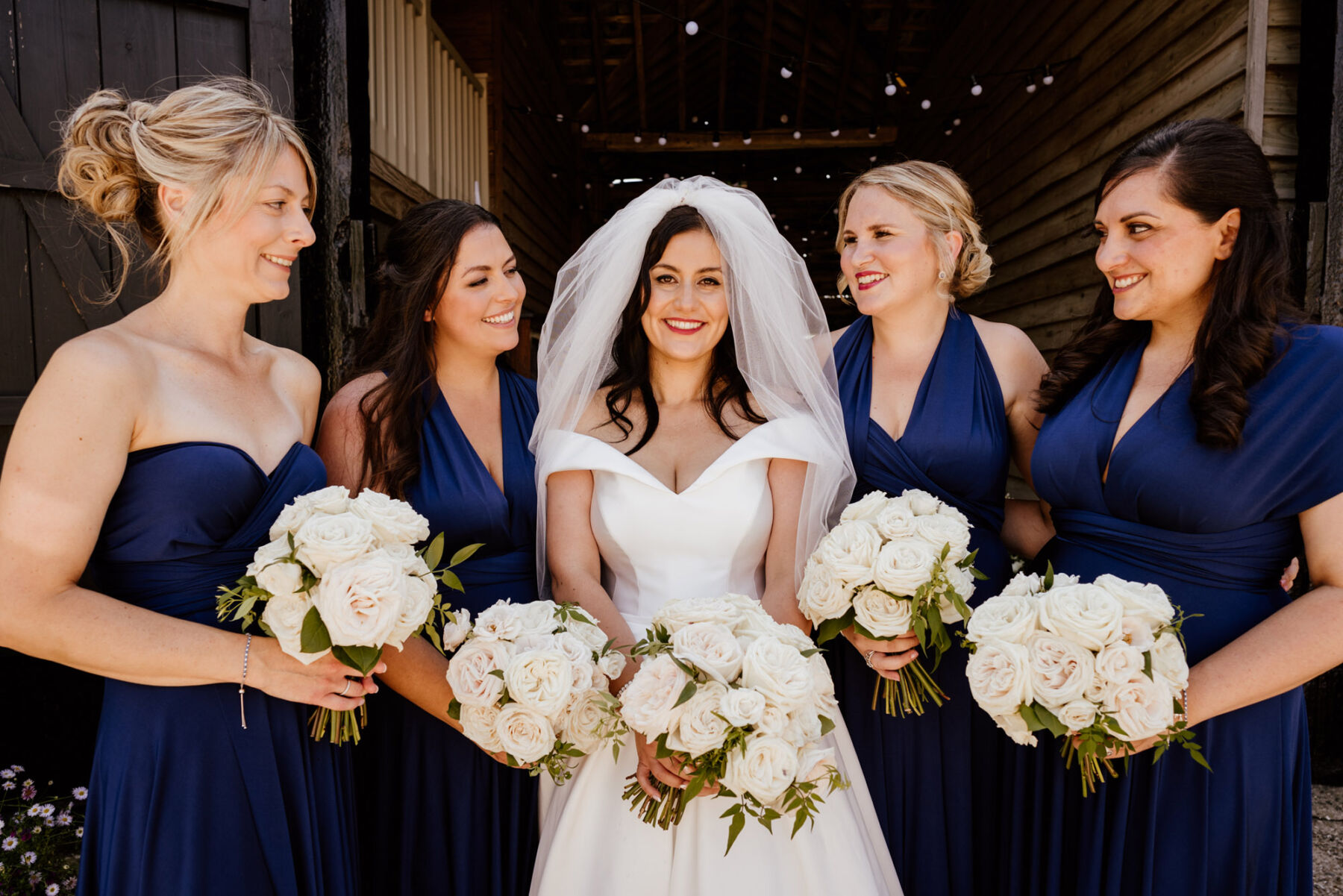 Bridesmaids wearing Navy Blue dresses & carrying bouquets of white roses. Bride in Suzanne Neville wedding dress.