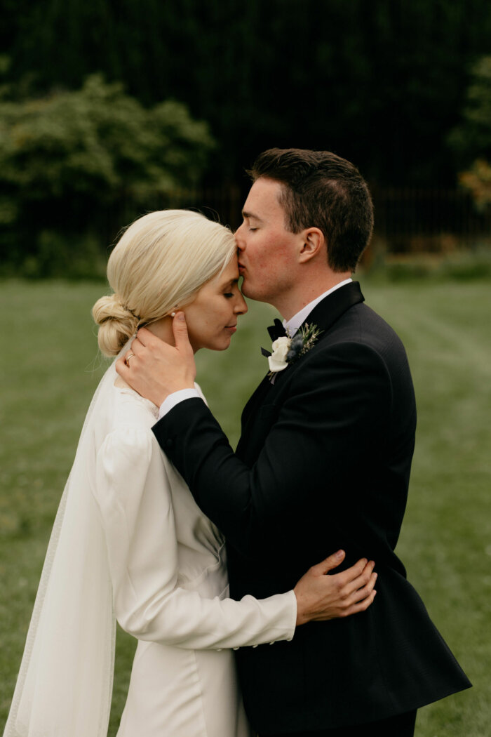 Groom in black tie gently embracing and kissing his bride. Bride has blonde hair and a low bun and wears a Rixo wedding dress and veil.