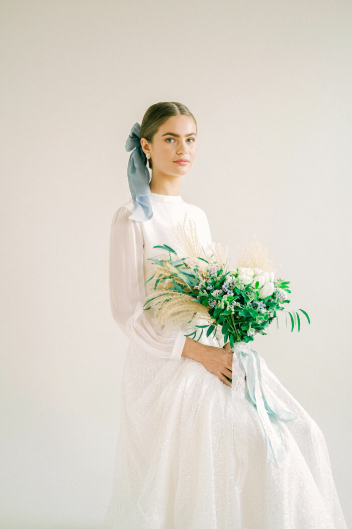 Bride wearing a Kate Beaumont wedding dress, blue silk bow in her hair & carrying a bouquet of green, cream and blue, with blue silk ribbons.