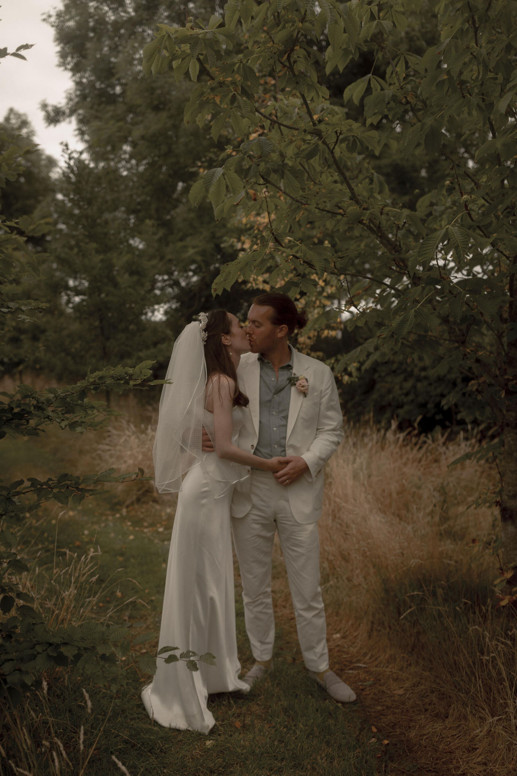 Bride & groom kissing under a tree. Galvan London wedding dress. Groom in white suit.