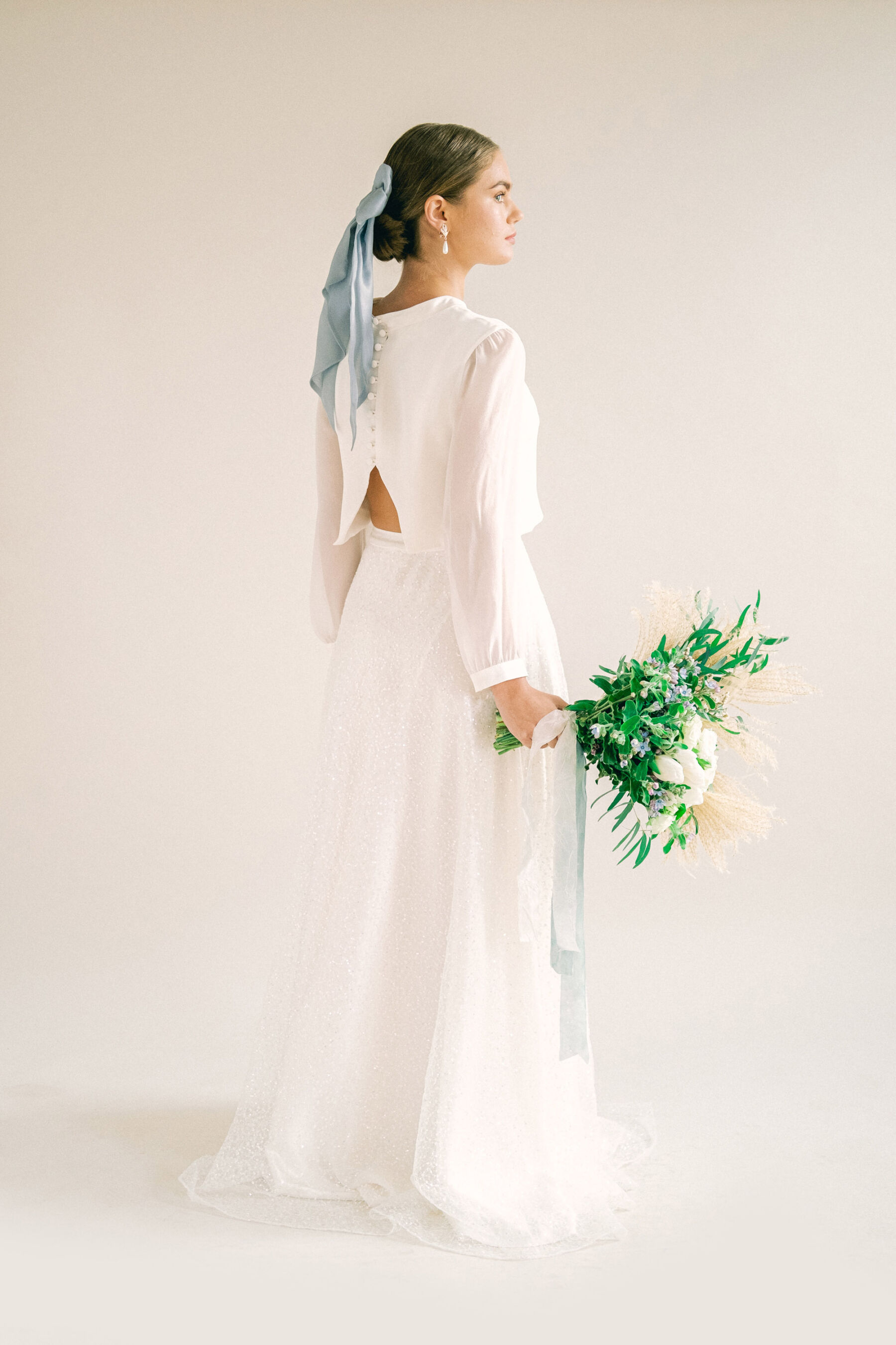 Bride wearing top and skirt bridal separates by Kate Halfpenny. Silk blue ribbon in her hair & carrying a wedding bouquet of mostly greenery with white and blue flowers.
