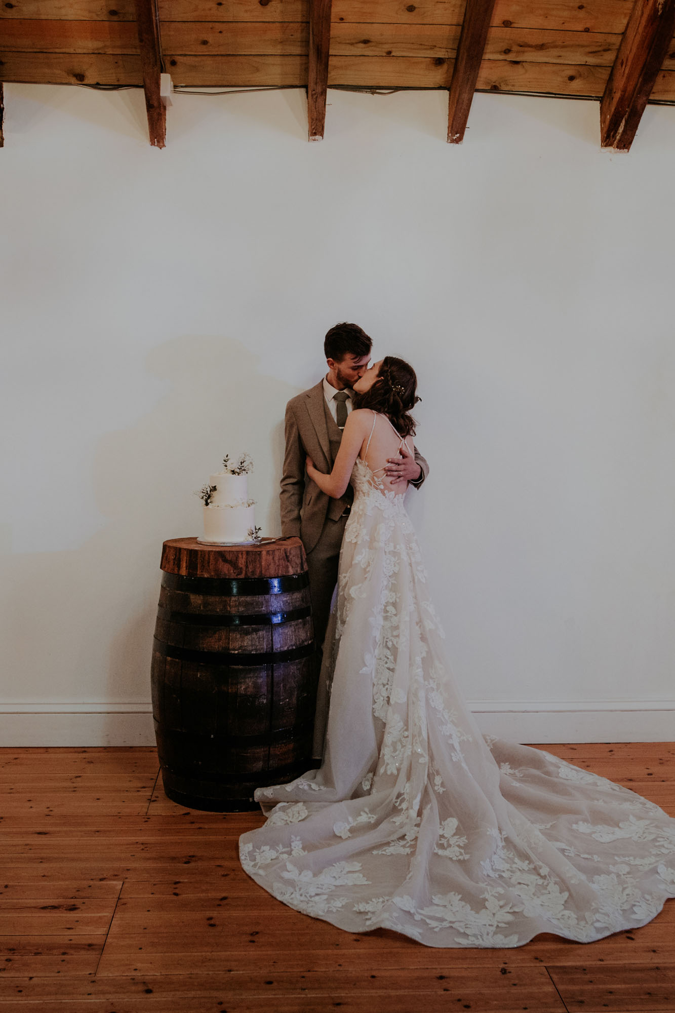 Bride in long lace dress cutting wedding cake on top of a wooden barrel 