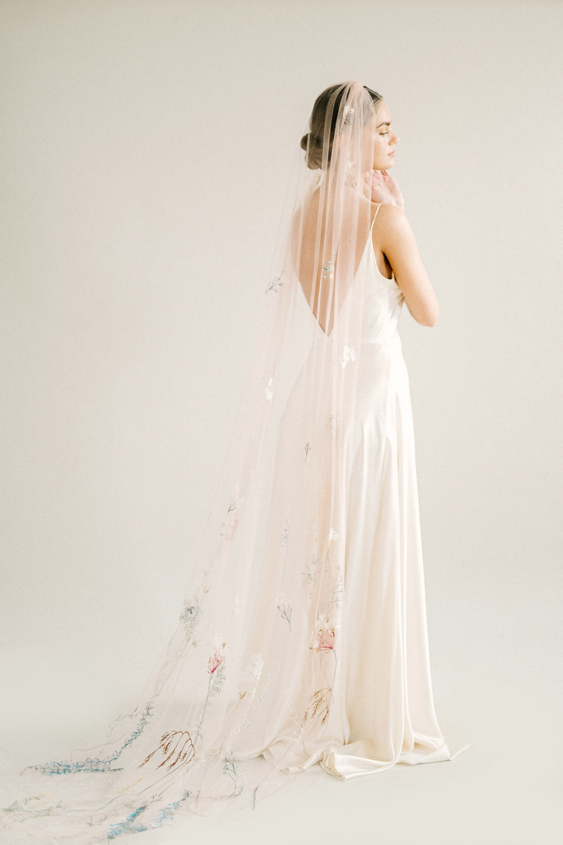 Bride with a low bun wearing a backless Kate Beaumont slip dress and Daisy Sheldon wedding veil embroidered with wild flowers