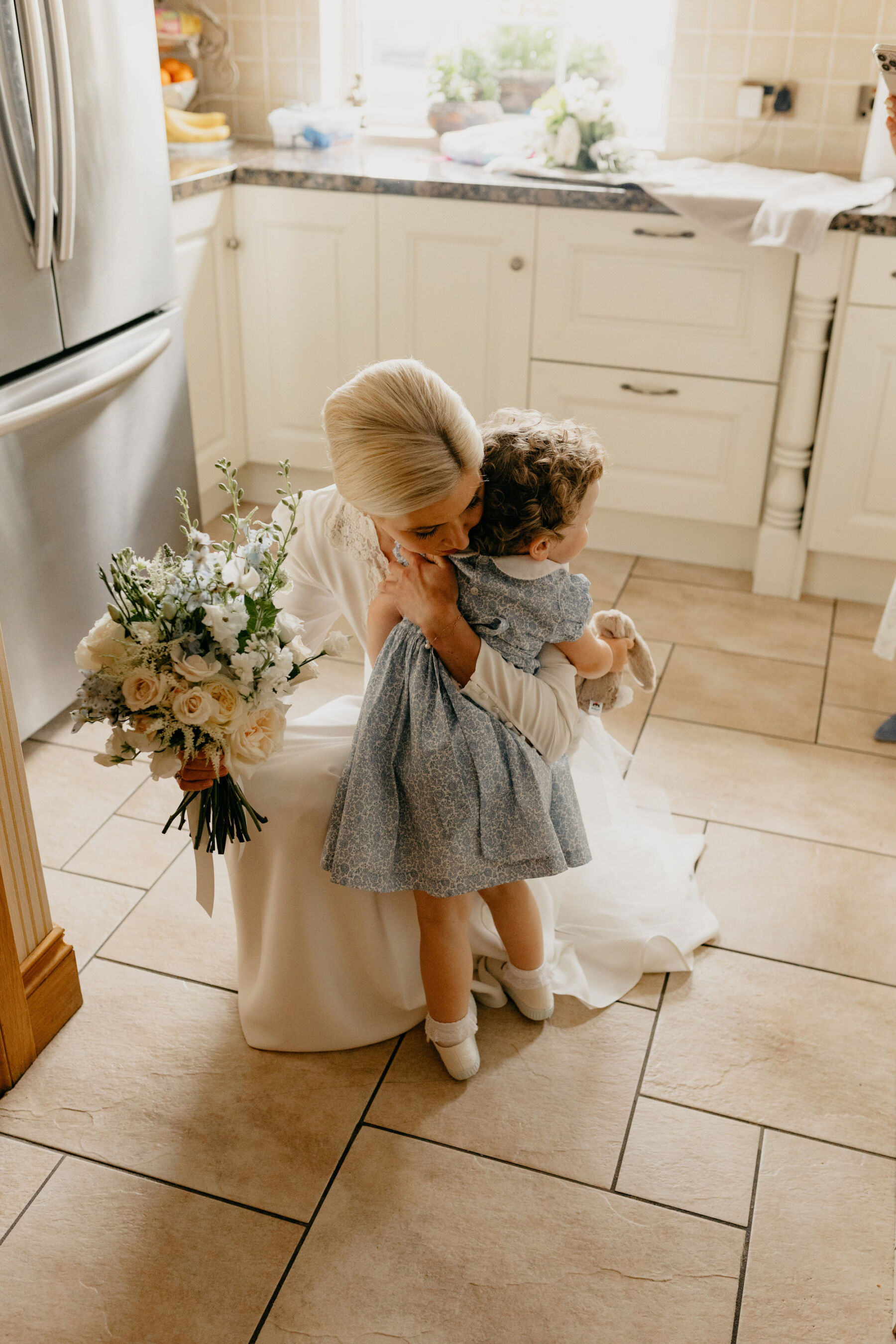 Bride in a Rixo wedding dress kneeling to embrace a little flowergirl in a pale blue dress.