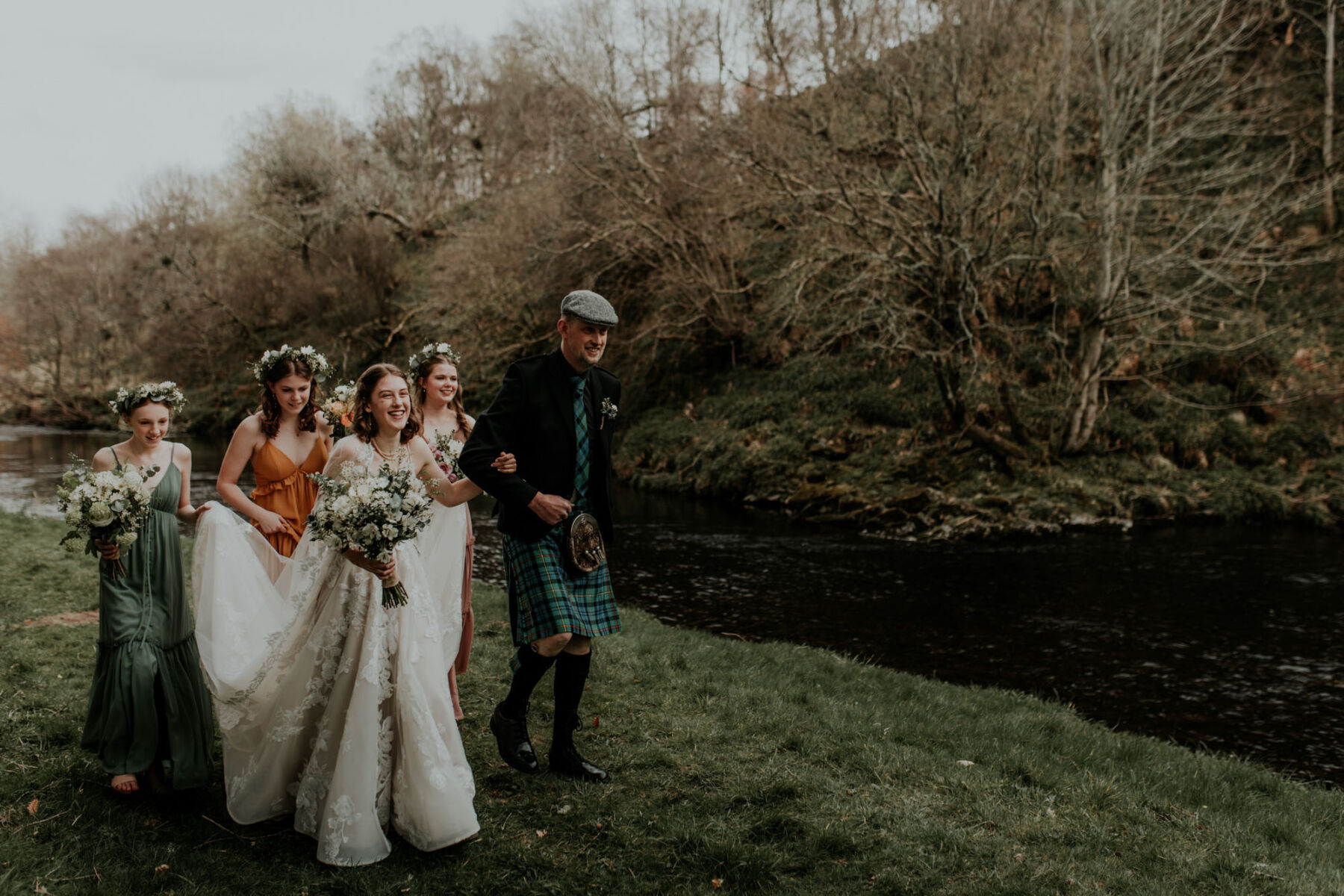 Bride making her way with dad in Kilt to her outdoor wedding ceremony at Aswanley. Bridesmaids in orange and green dresses carrying the bride's dress train.