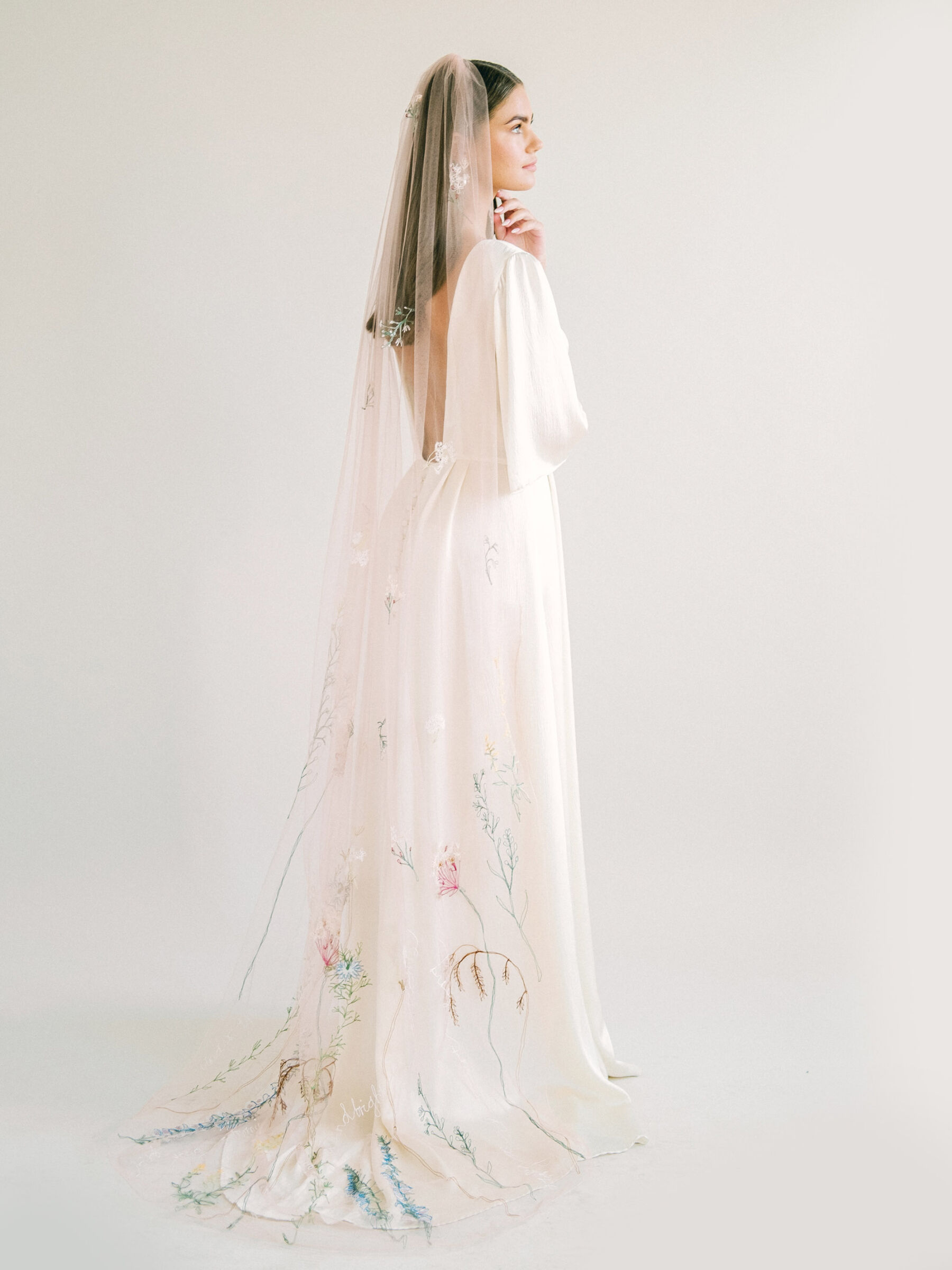 Bride standing with her back to the camera, wearing a low-back dress by Kate Beaumont & chapel length, long wedding veil with floral embroidery by Daisy Sheldon.