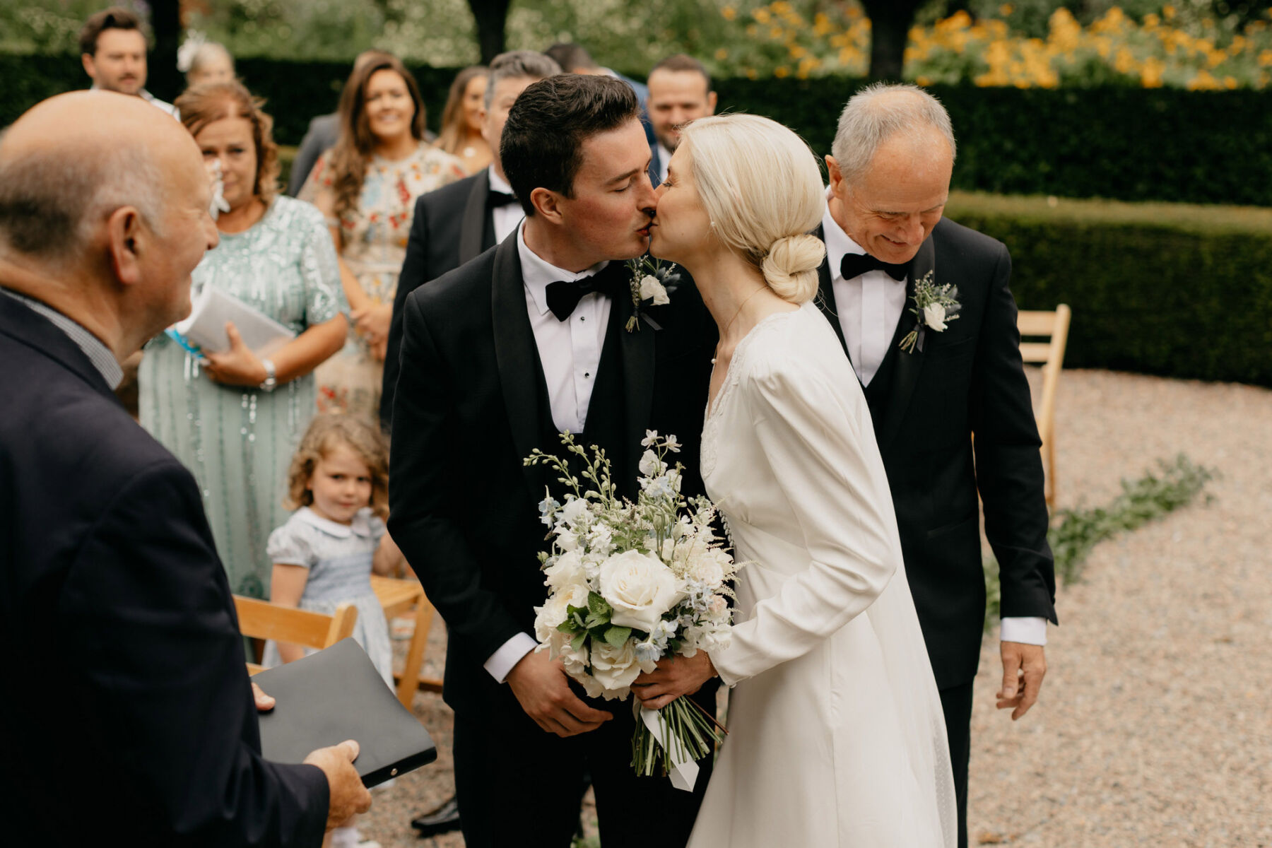 Black tie wedding, groom kissing his bride in Rixo wedding dress. Bride has a low bun and carries a white wedding bouquet. Outdoor wedding at Larchfield Estate in Co Down Northern Ireland.