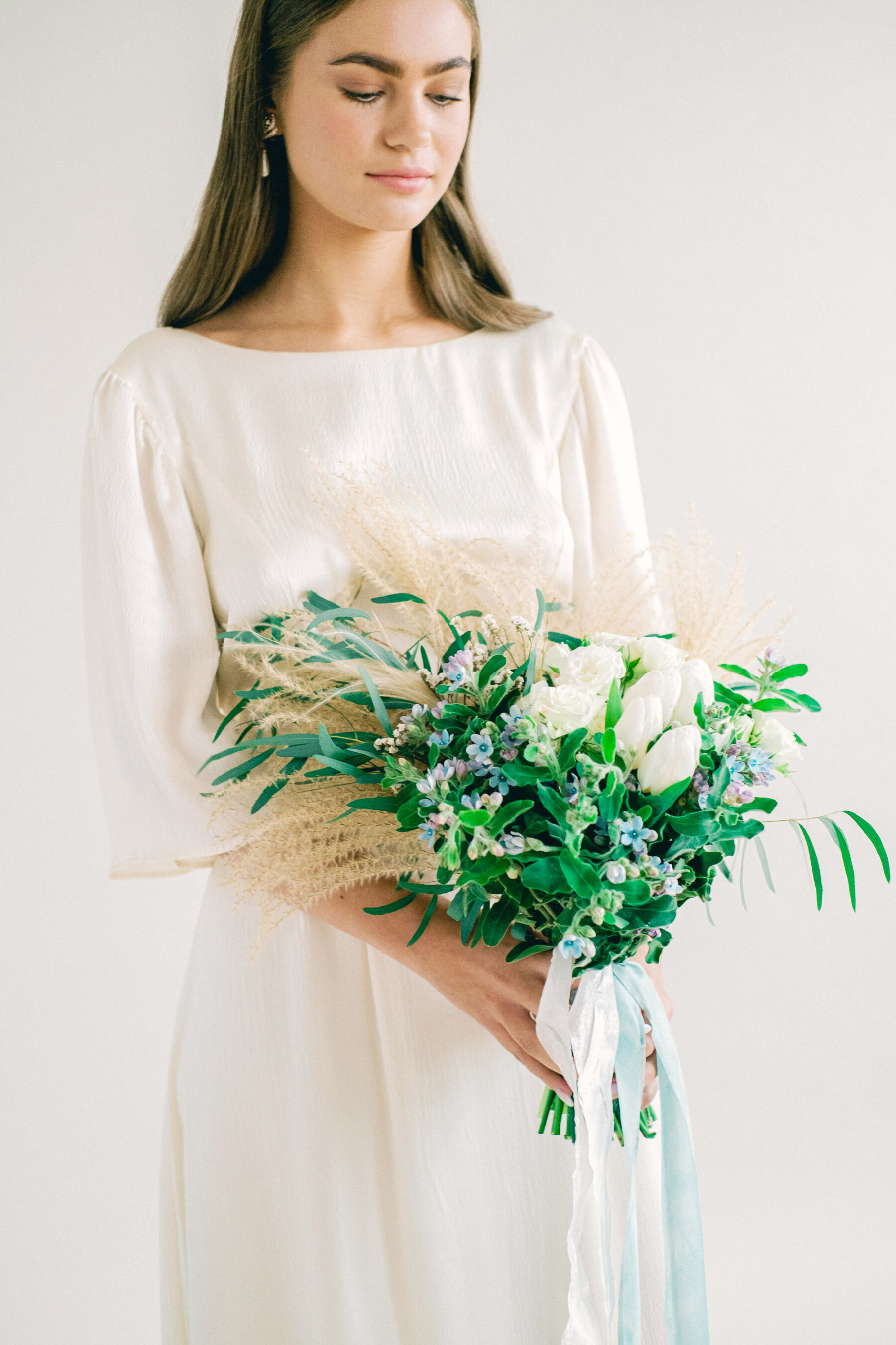 Bride wearing a Kate Beaumont wedding dress & carrying an elegant bouquet with greenery & blue flowers & long silk cream & blue ribbons.
