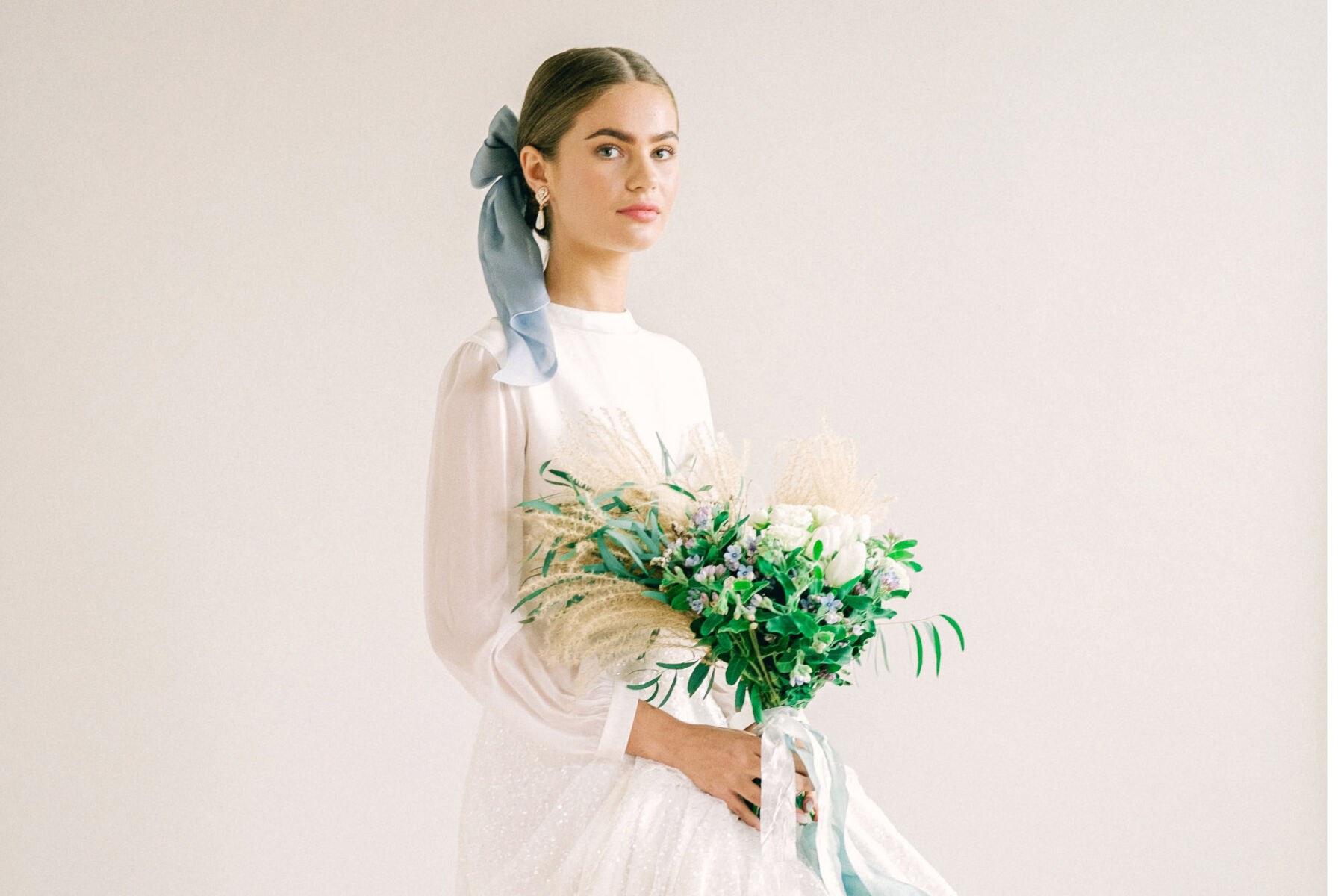 Bride with hair tied back in to a pale blue silk ribbon, holding a bouquet of blue flowers and grasses and wearing a Kate Beaumont wedding dress.