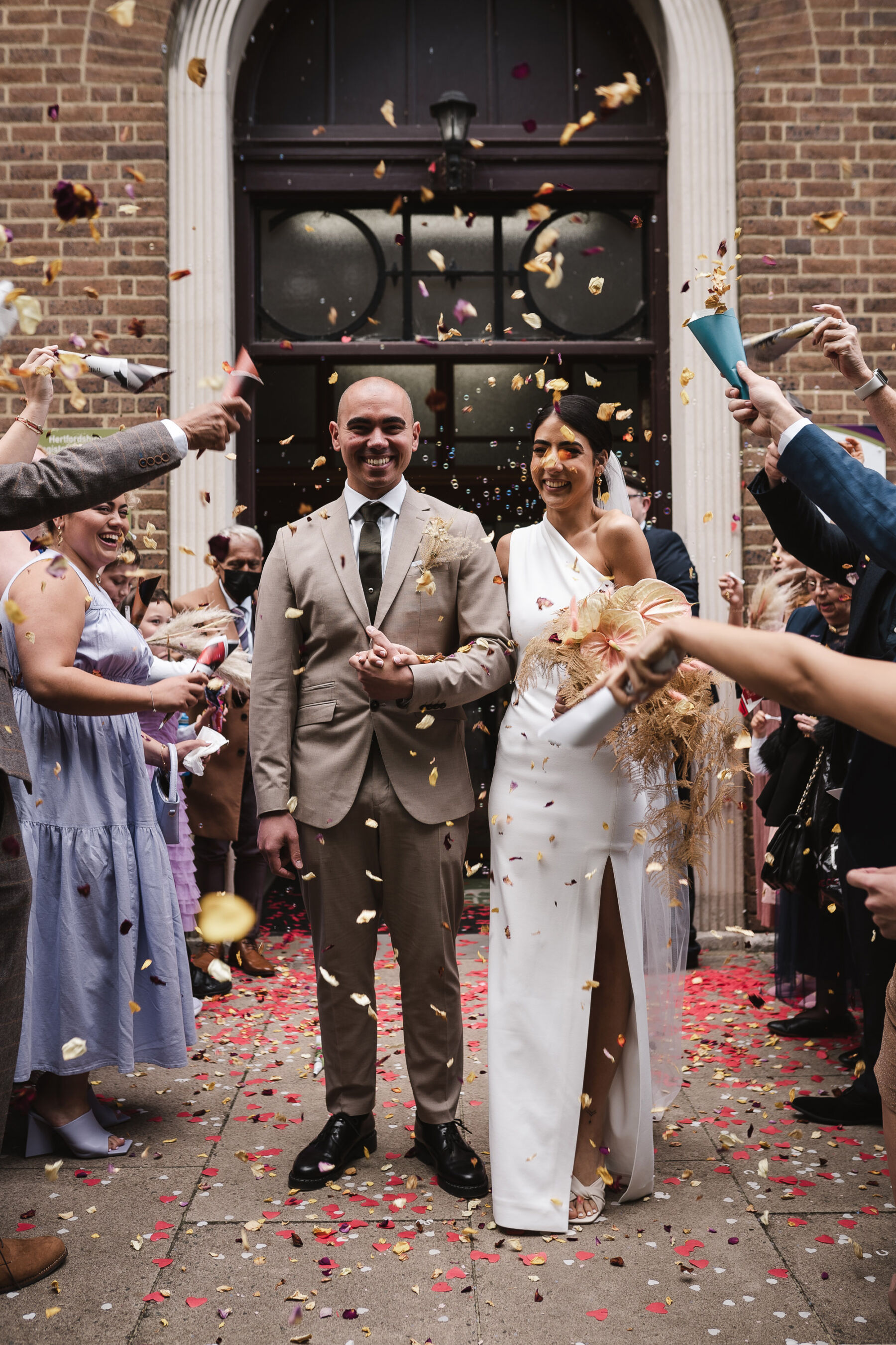 Confetti outside the registry office. Groom in pale beige suit. Bride in Solace London one shoulder asymmetrical wedding dress. By Karolina Photography.