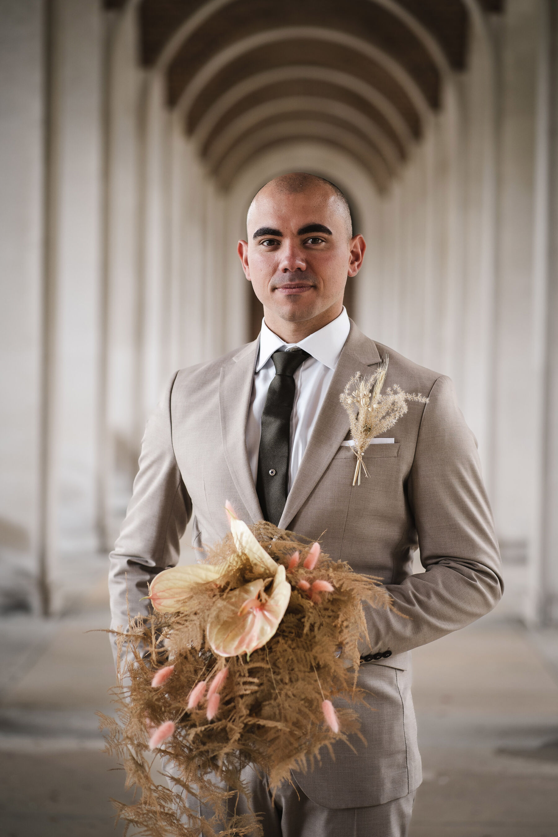 Groom in light beige suit holding a dried flower & fern bouquet. By Karolina Photography.