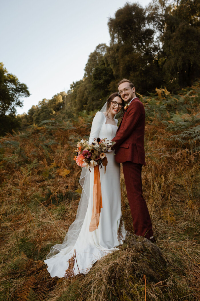 Bride wears Amber by Suzanne Neville. Groom in dark red suit. Bride carries Autumn wedding bouquet.