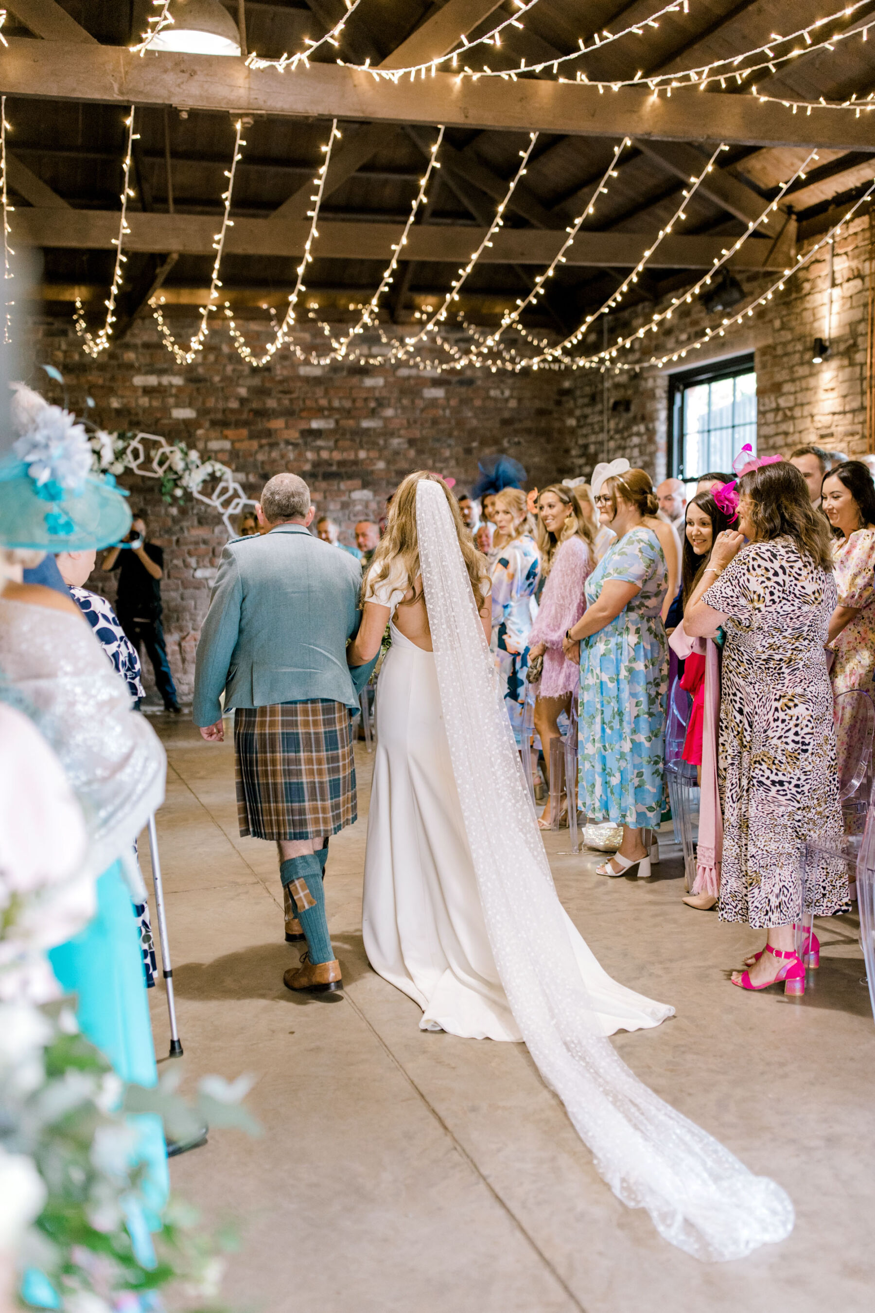 Bride wearing a Sarah Seven backless wedding dress, walking beneath a canopy of fairylights at Glasgow Engine Works industrial wedding venue