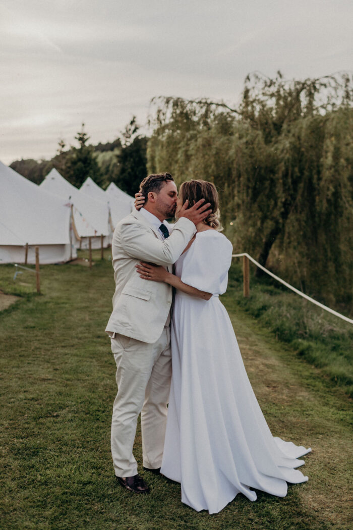 Groom in pale suit kissing bride in & For Love dress, couple standing by tents on a farm.