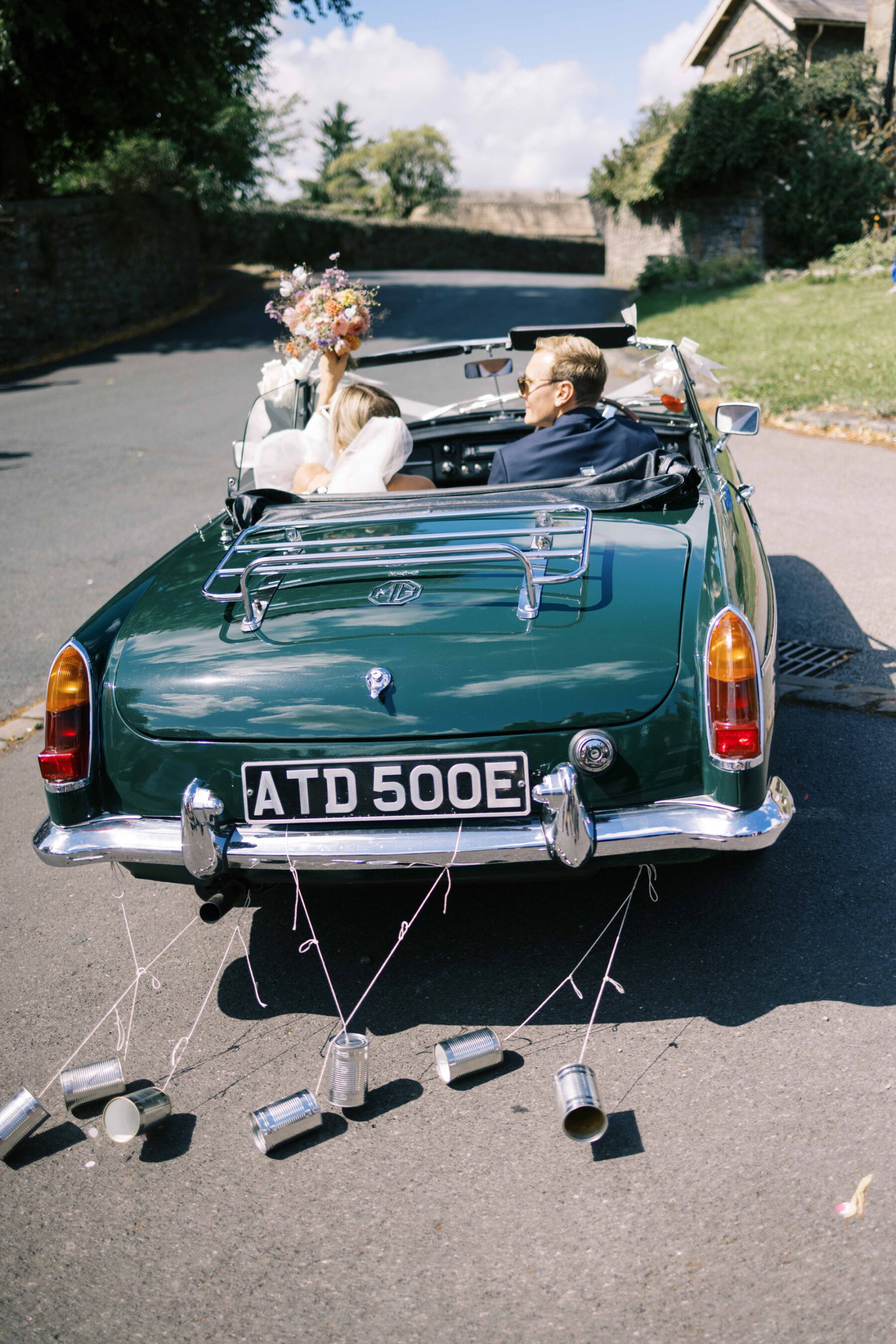 Bride and groom in an open top vintage car with wedding tin cans trailing at the back. Image by Emmy Shoots.