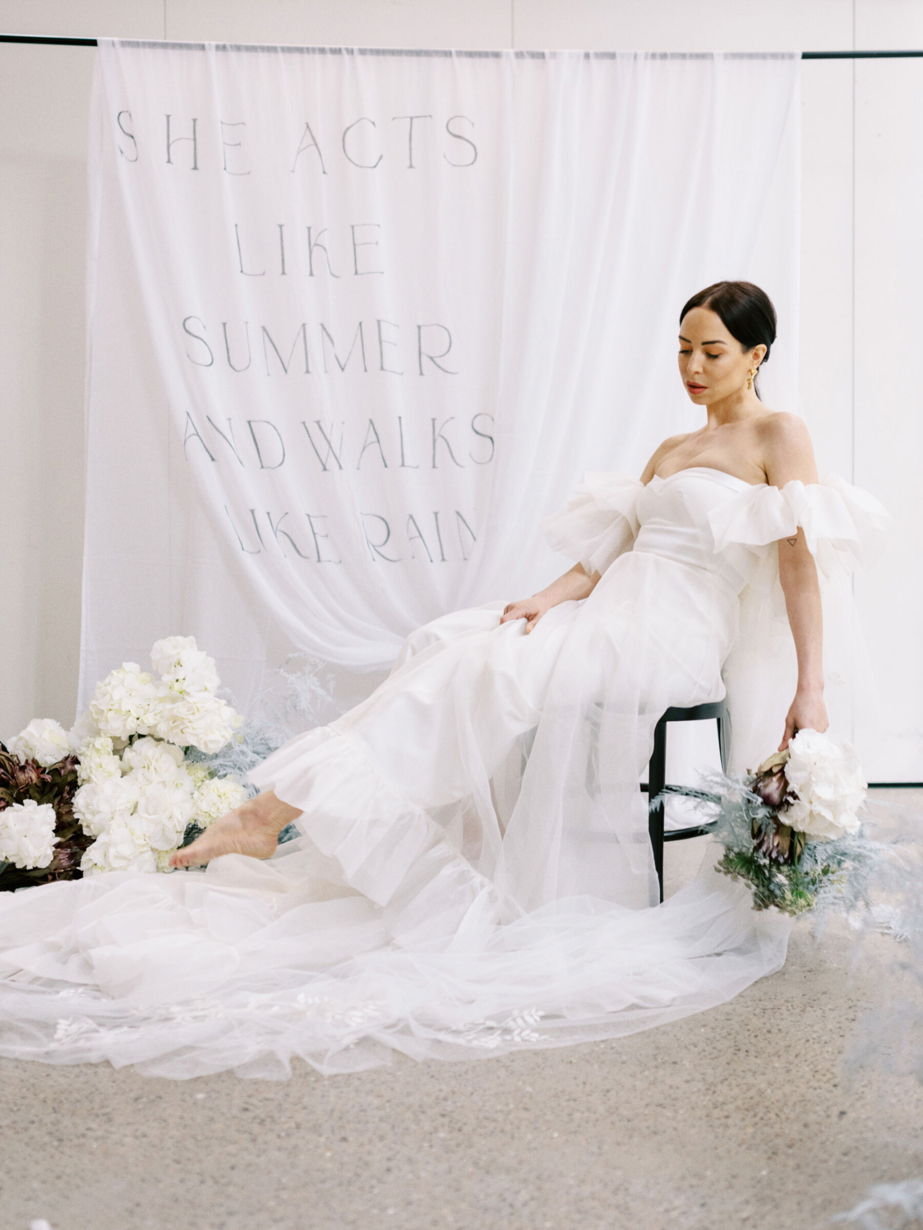 Barefoot bride in Halfpenny London sitting back on a chair with hydrangea and fabric hanging nearby with romantic words printed on it