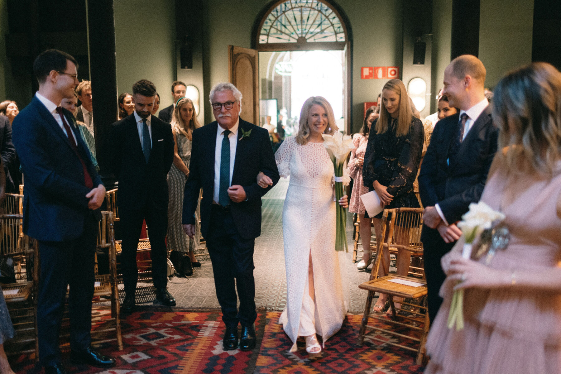 Bride being walked down the aisle by her father, she wears Immacle and carries a bouquet of white Hippeastrum. Rebecca Rees Photography. 