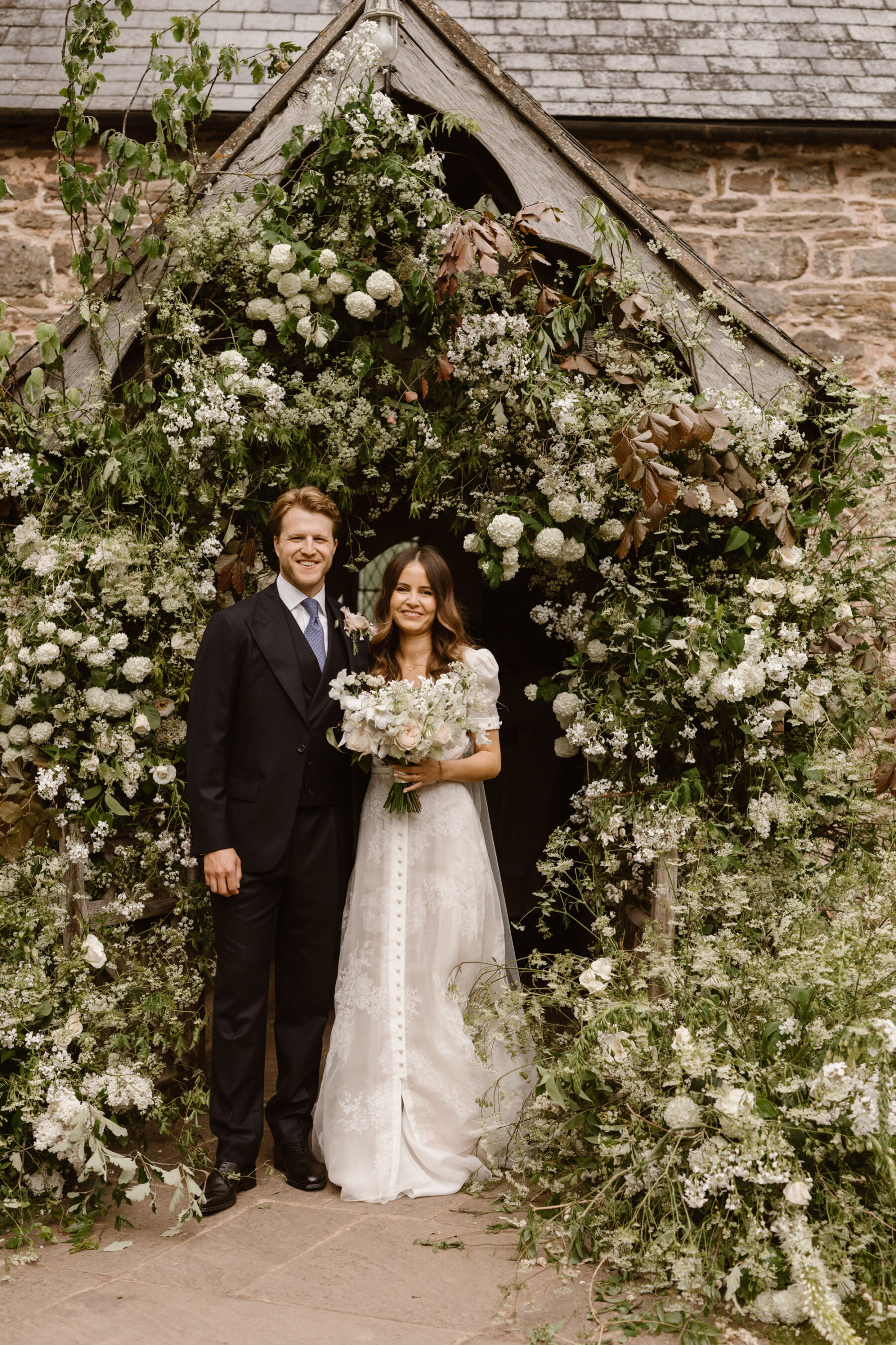Bride and groom surrounded by a floral arch surrounding the entrance to Dewsall Church. Bride weas J Andreatta wedding dress from Amare. Agnes Black Photography.