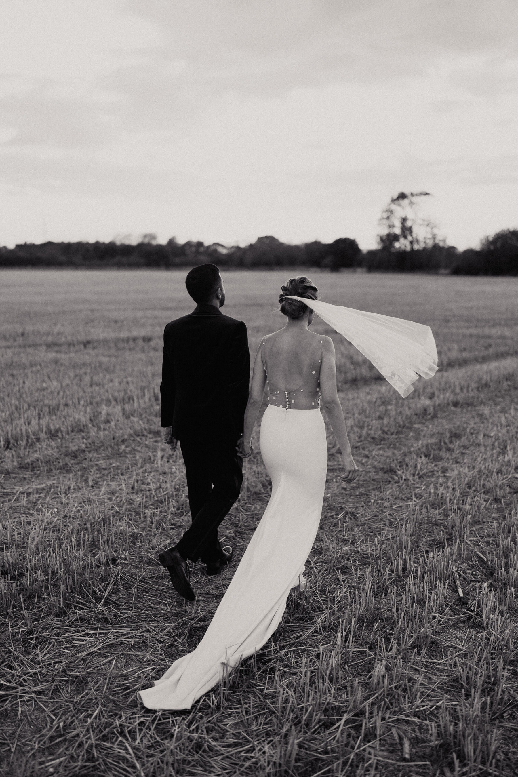 Black and white image of glamorous bride and groom in a field. Bride's short veil is flying away in the breeze.