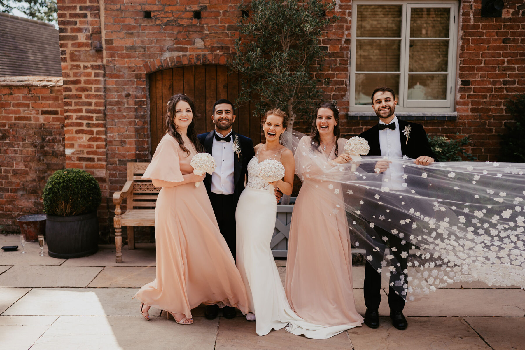 Bridesmaids in peach dresses, groom in tuxedo, bride in a long flyaway wedding veil.