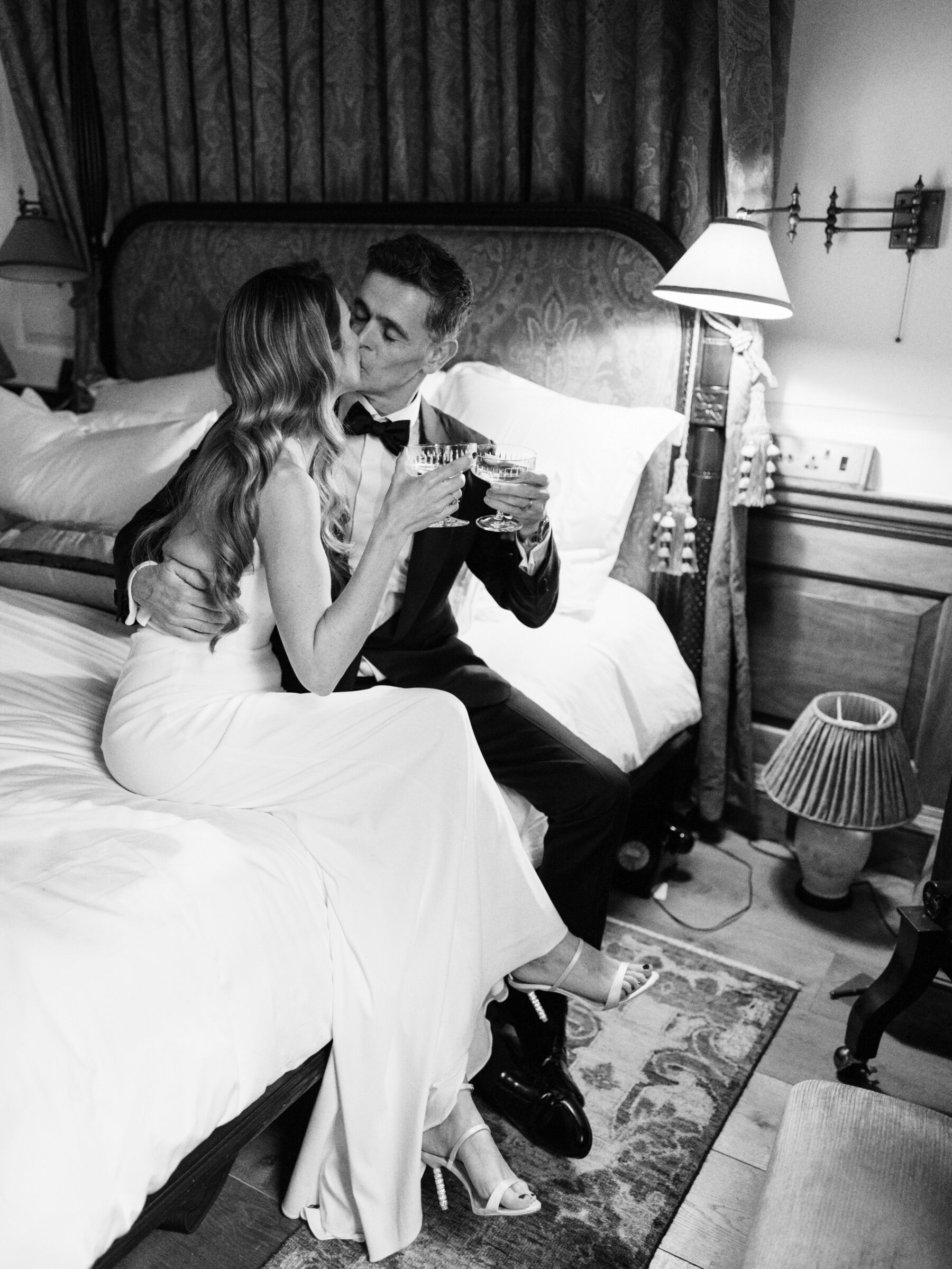 Bride and groom embracing on the bed at The Ned, London and drinking champagne. Kernwell Photography.
