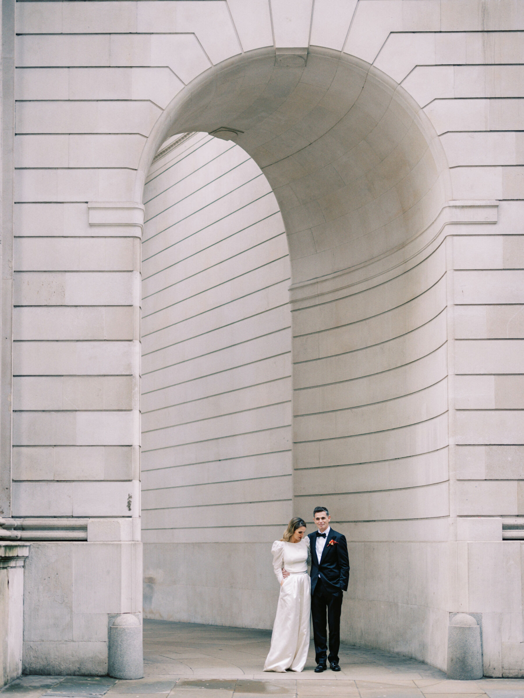 Bride and groom standing by The Ned, London. Bride wears modern wide leg bridal trousers by Halfpenny London. Kernwell Photography.