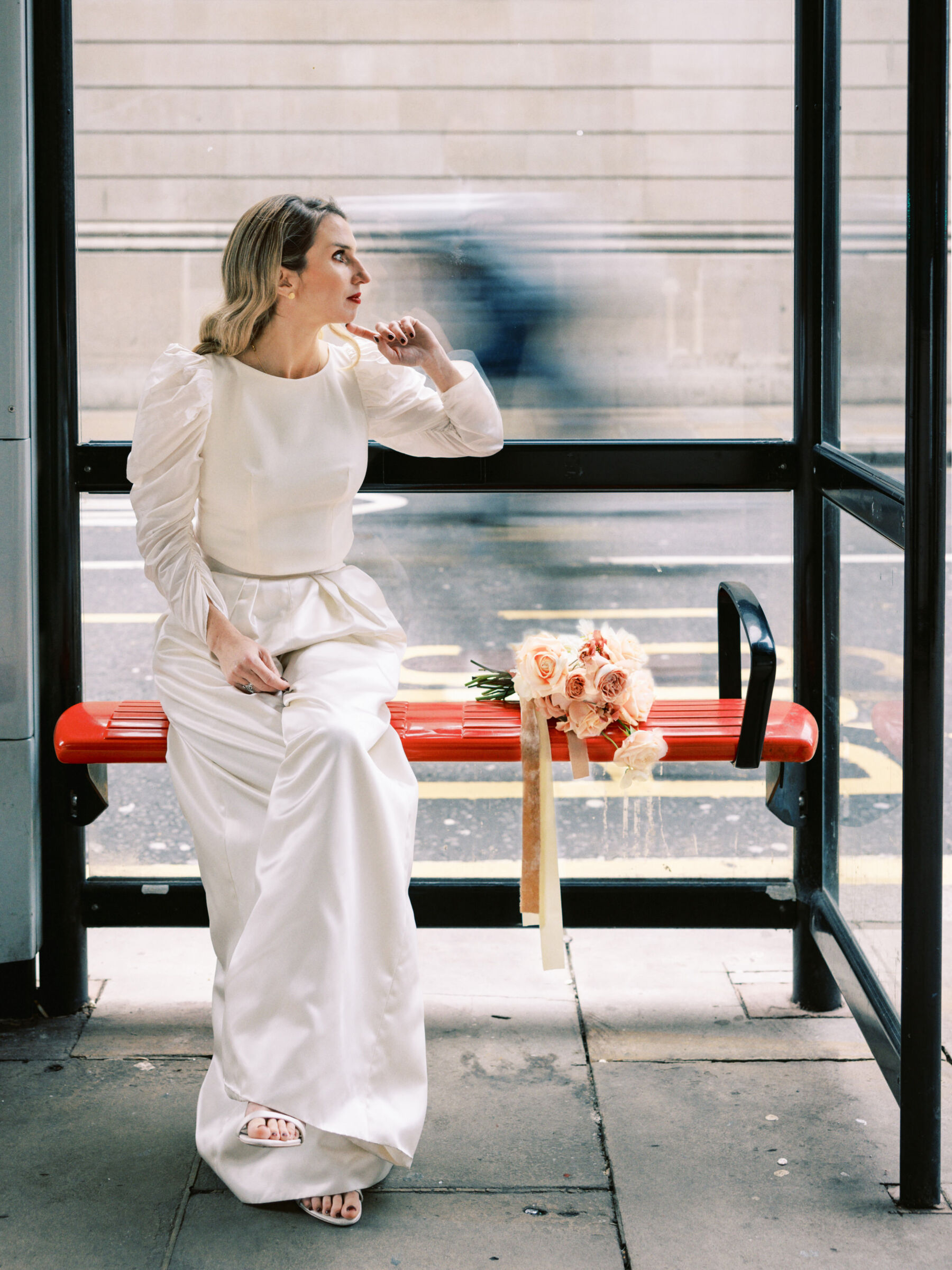 Bride sat at a bus stop in London, wearing modern wide leg bridal trousers by Halfpenny London. Kernwell Photography.