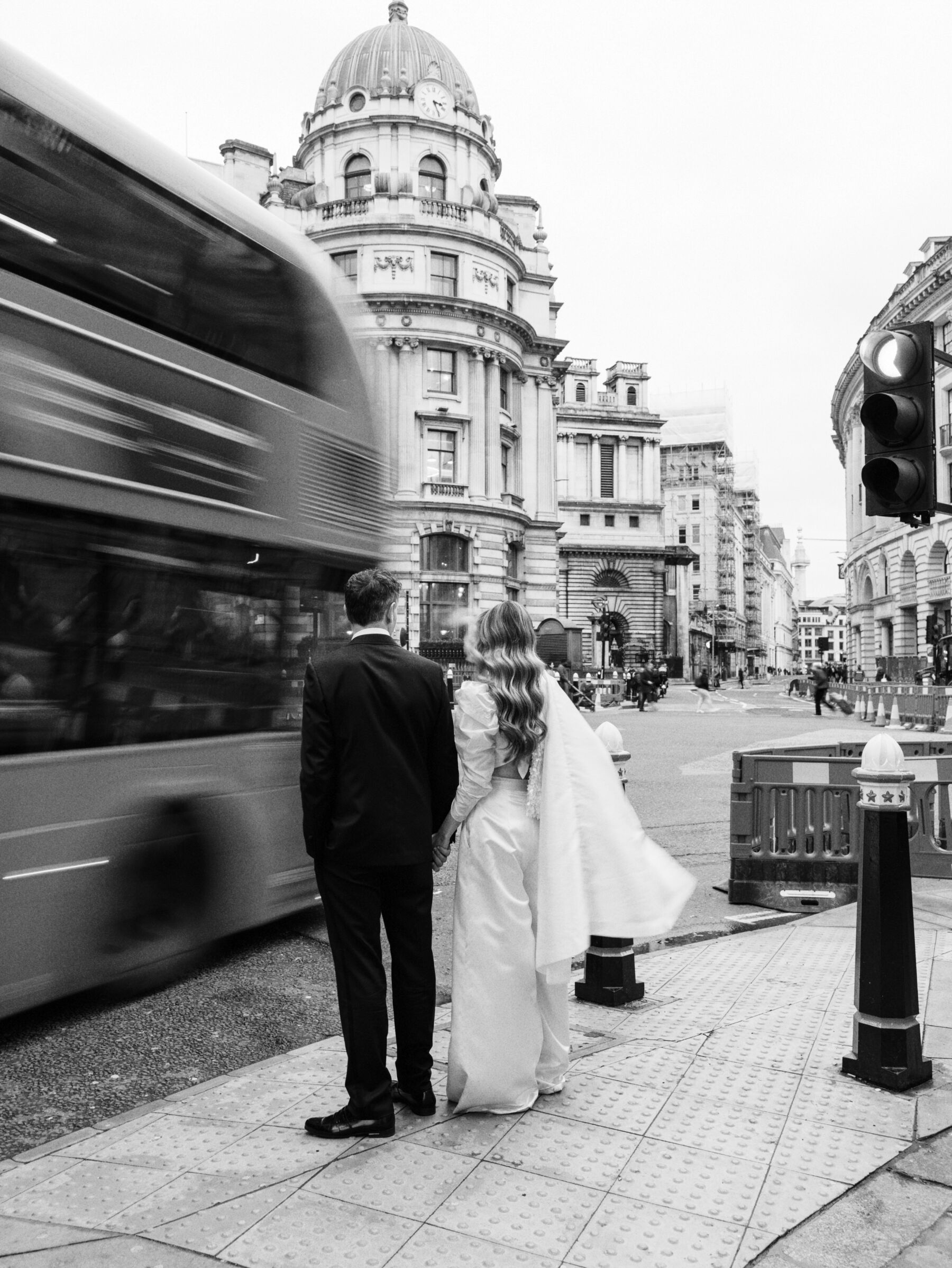 Bride and groom waiting for a bus to pass on the streets of London. Bride wears modern wide leg bridal trousers by Halfpenny London. Kernwell Photography.