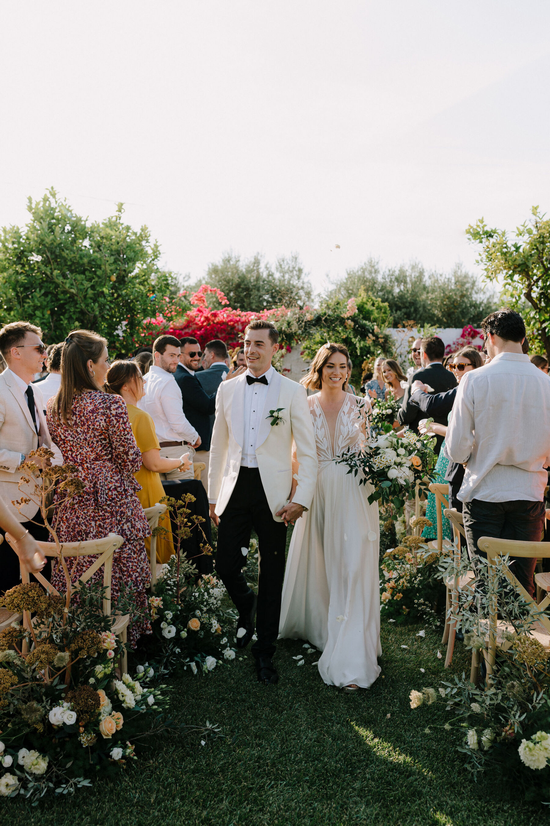 Bride in Margaux Tardits dress and groom in white tuxedo leaving their outdoor wedding ceremony in Puglia, Italy.