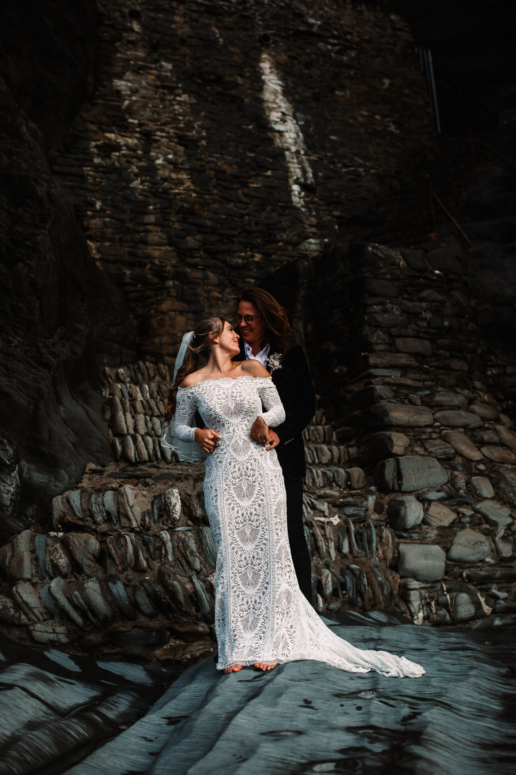 Bride and groom stand on the rocks at Tunnels Beaches wedding venue in Devon. Bride wears Grace Loves Lace.