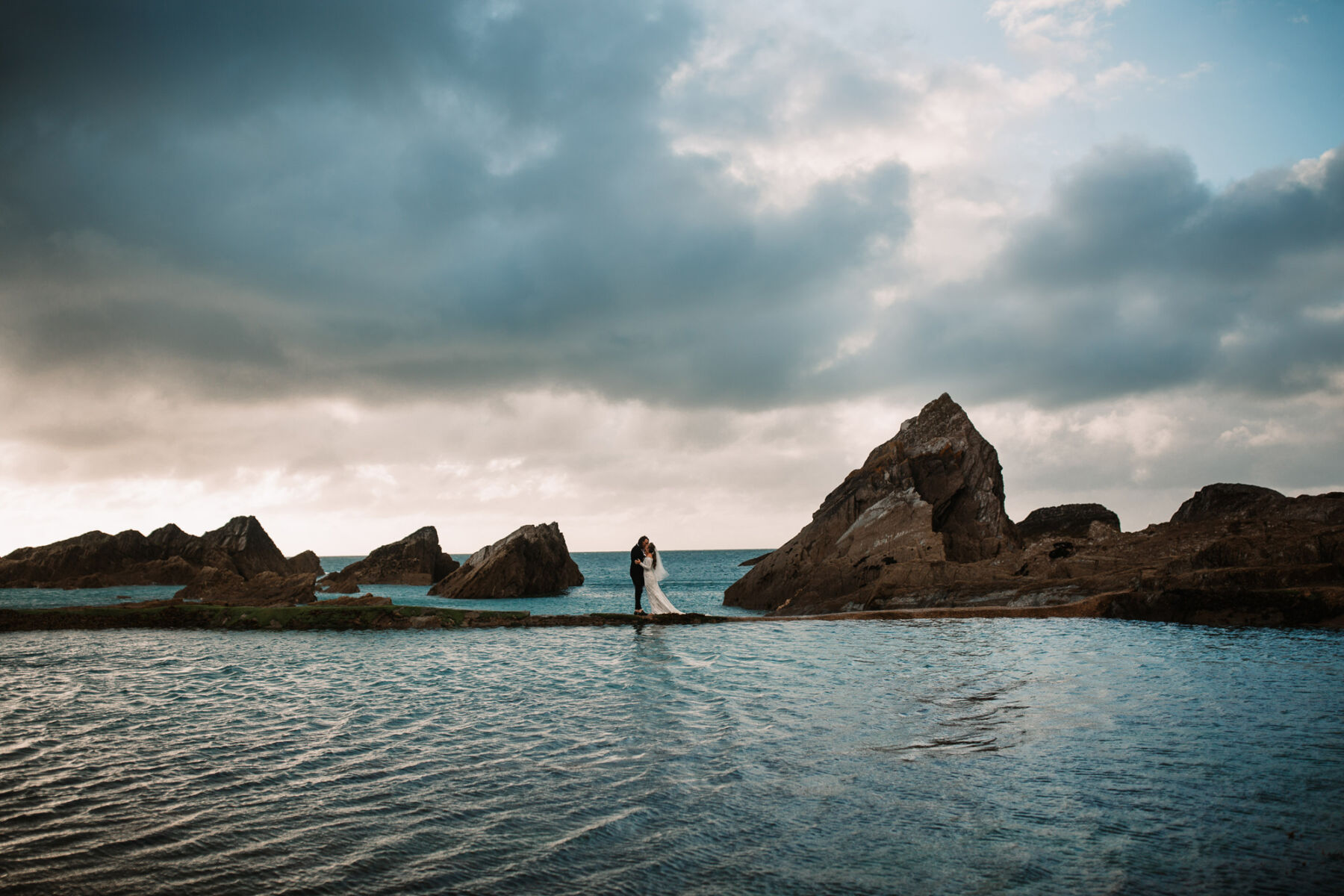 A Devon Wedding with a Sea View for Georgie in Grace Loves Lace 46 Bride and groom stand on the rocks at Tunnels Beaches wedding venue in Devon. Bride wears Grace Loves Lace.