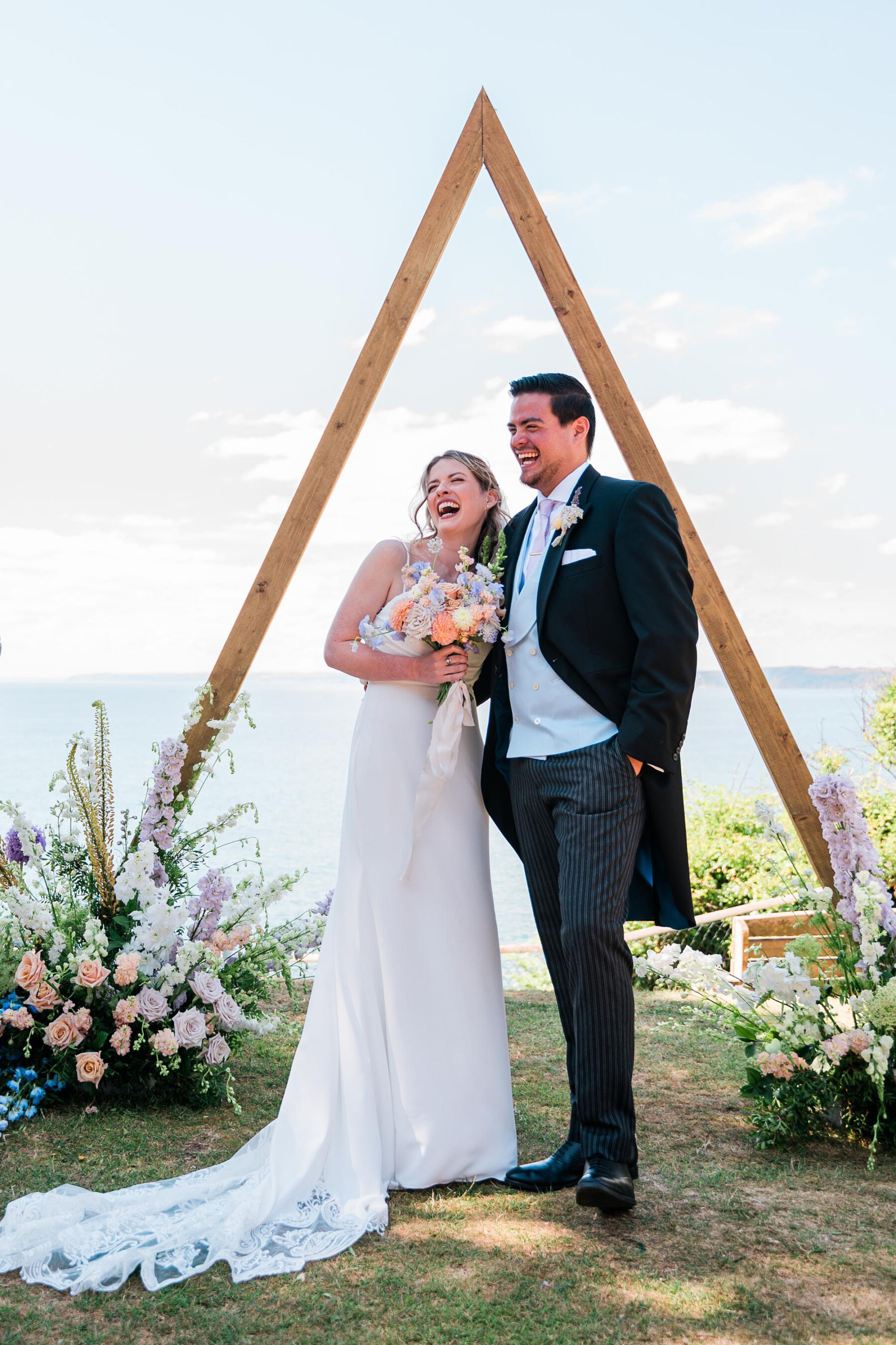 Sea view wedding in Cornwall. Bride and groom stood next to a triangle wooden ceremony backdrop with summer flowers at it's base.