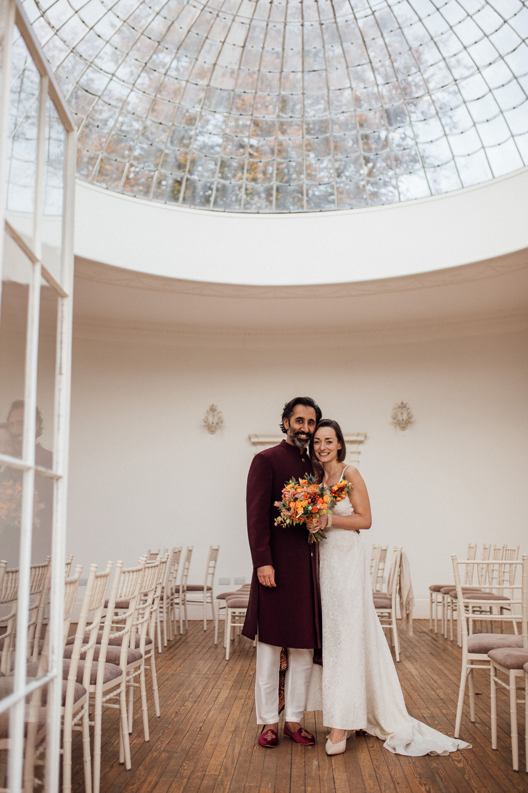 Bride wearing a 1950's wedding dress standing with her Asian groom in the Orangery at Barton Hall.