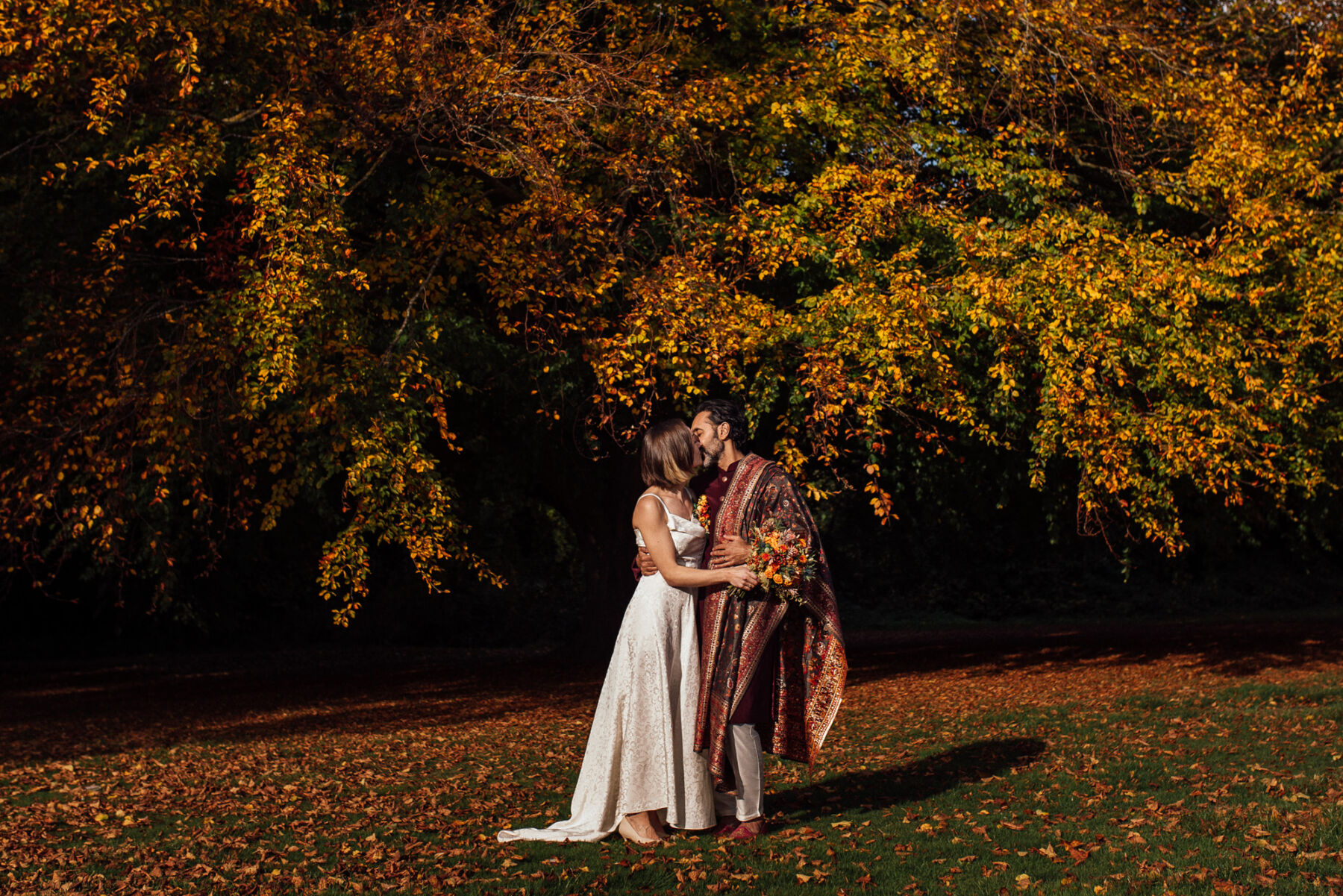 Mixed race bride and groom standing in Autumn leaves that have fallen from the trees at Barton Hall.