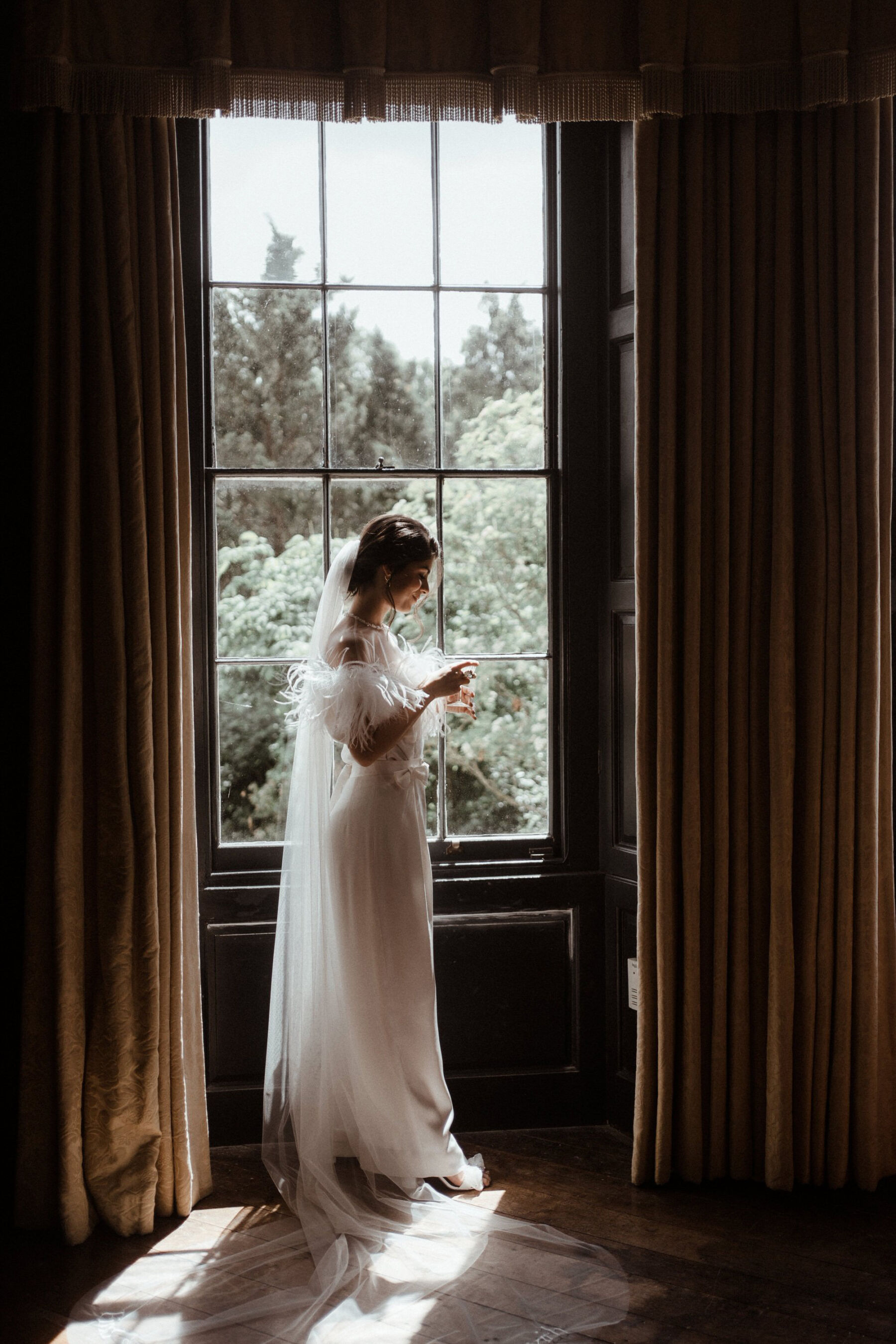 Bride standing in a window at Elmore Court, wearing Wilden London trousers and embroidered veil by Rebecca Anne Designs.