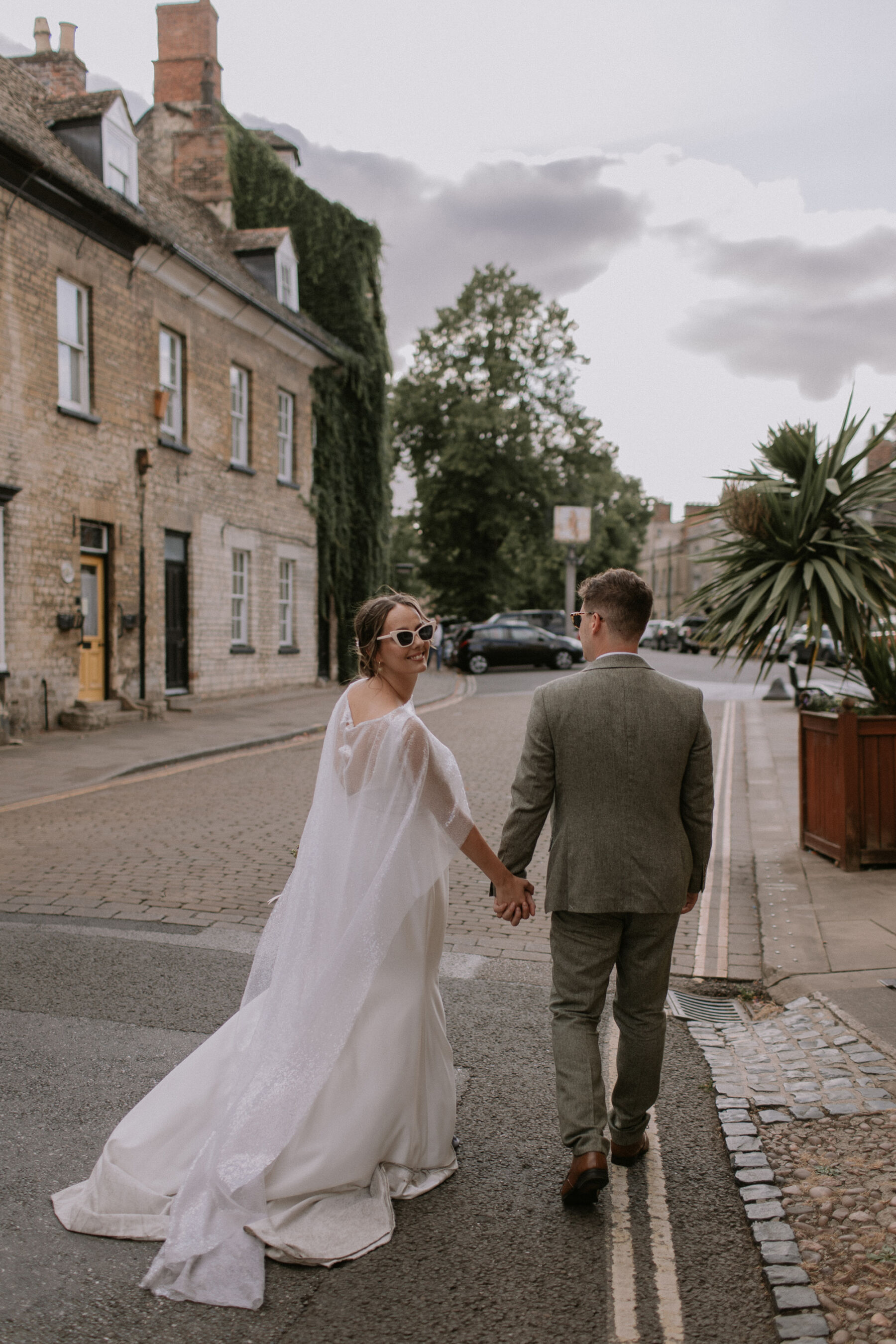 Bride in sunglasses and Charlie Brear dress and sequin cape. Groom in Moss Bros suit.