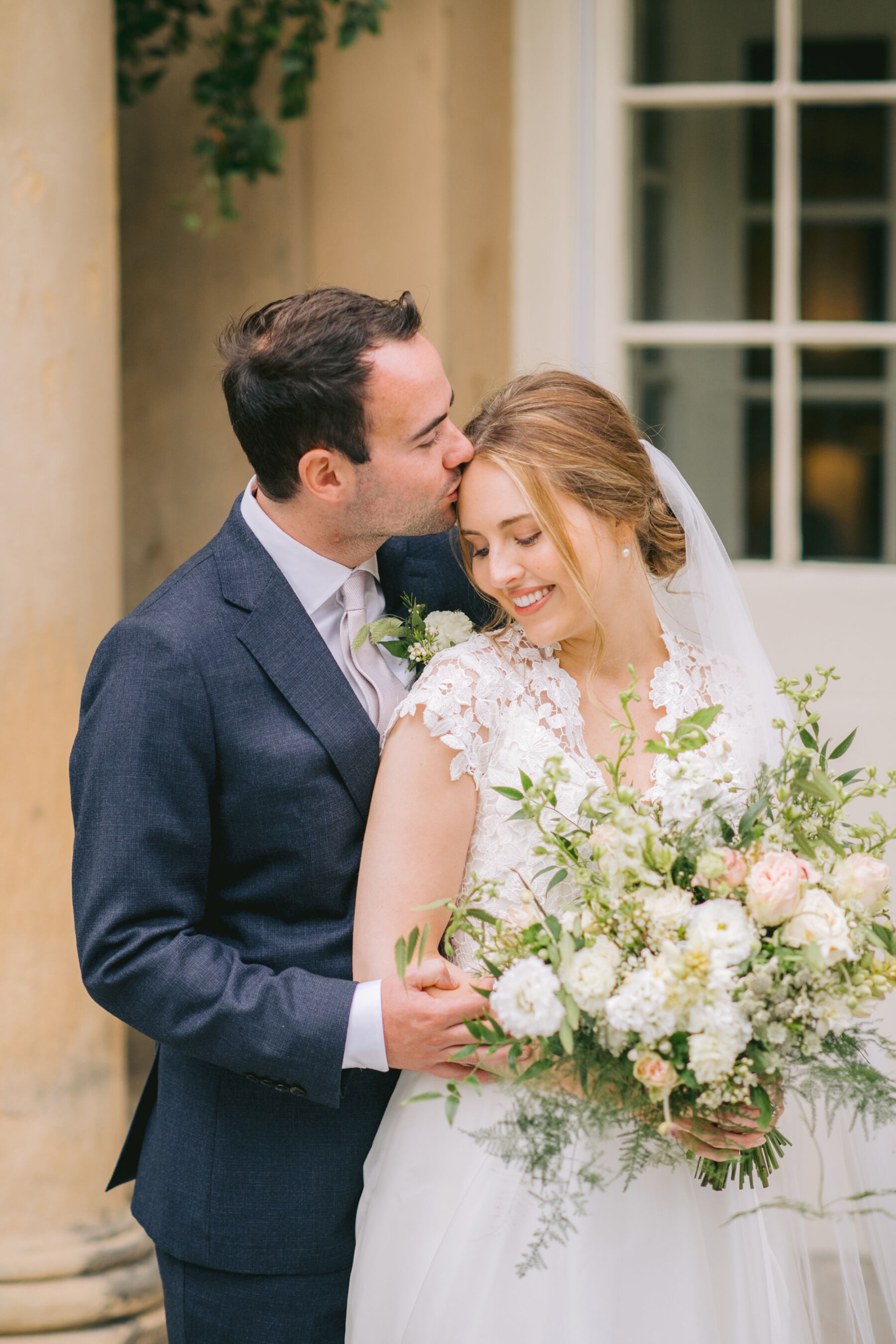 Groom kissing his bride in Caroline Castigliano wedding dress, carrying a beautiful seasonal summer wedding flowers bouquet.