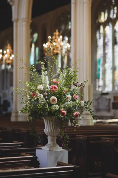 A single garden urn full of roses, jasmine and tall delphinium catching the light as it filters through the stained glass windows of St Margarets Church, Westminster Abbey.