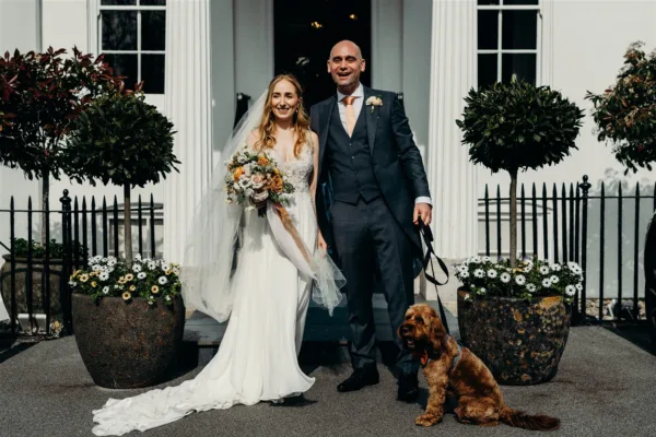A bride & groom stand outside a doorway, the bride is holding her bouquet, as ribbons in matching colours billow in the wind. The groom is holding their pet dog.
