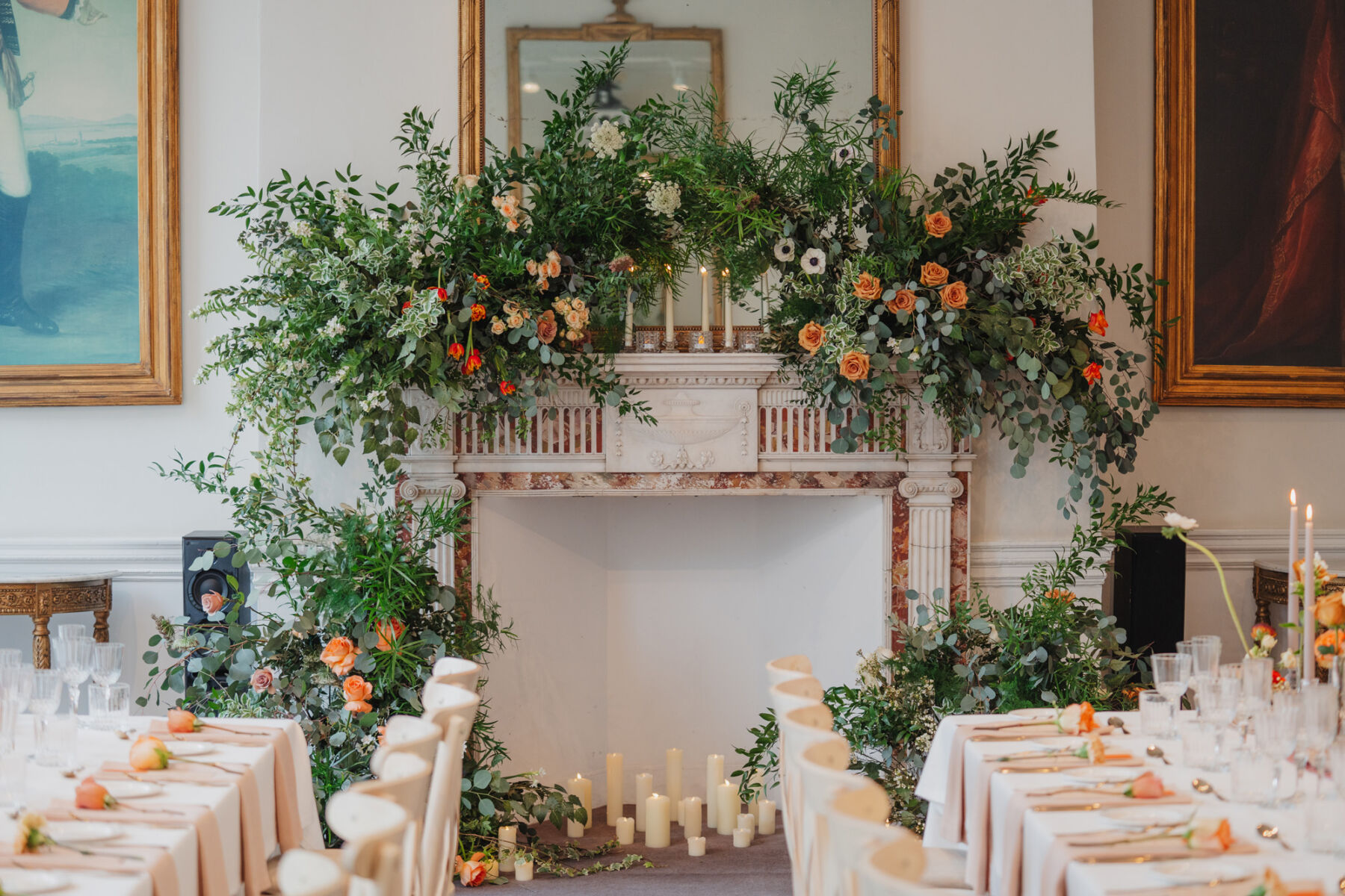 Sprawling foliage and orange flowers decorating the fireplace and mantlepiece at 41 Portland Place London. Candles of various height sit inside the fireplace. 