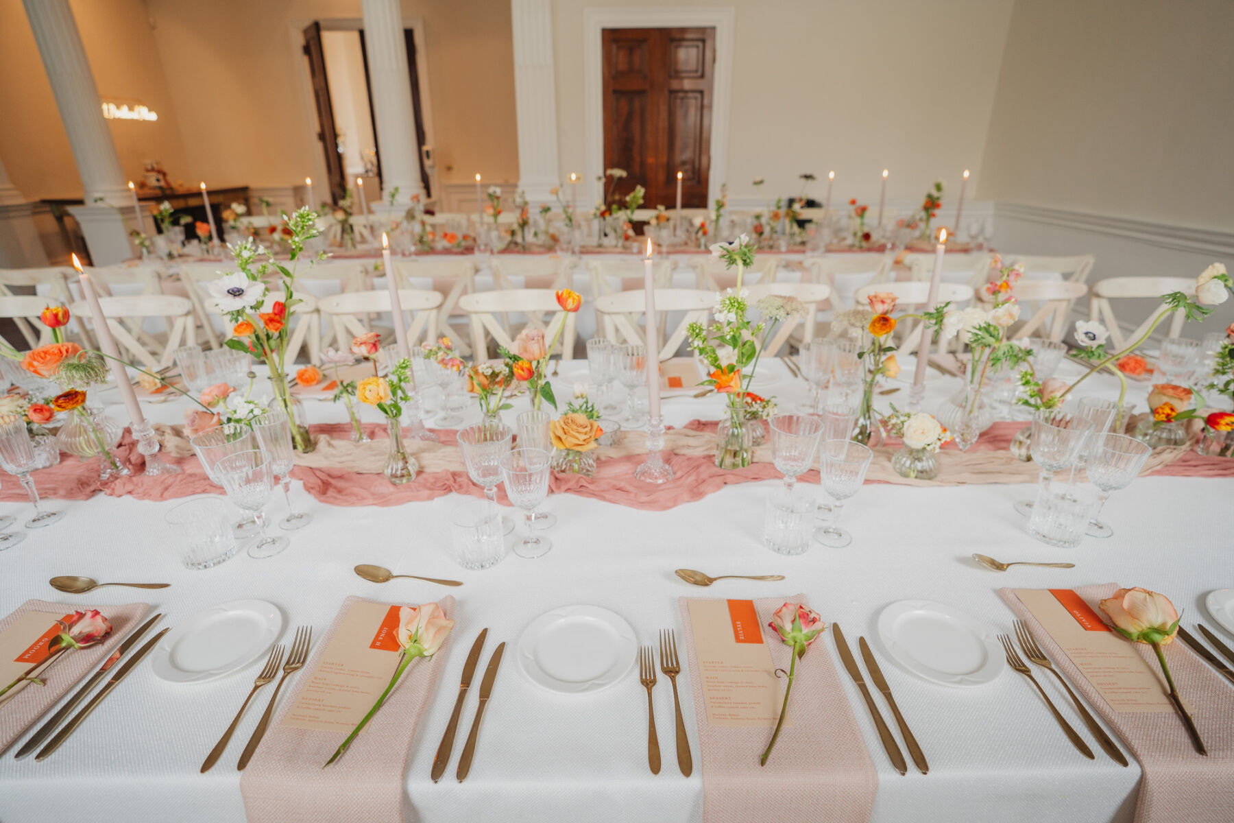 Wedding reception setup on long tables with white linen, cream taper candles and orange wedding flowers and menus. A single stem rose decorates each setting.