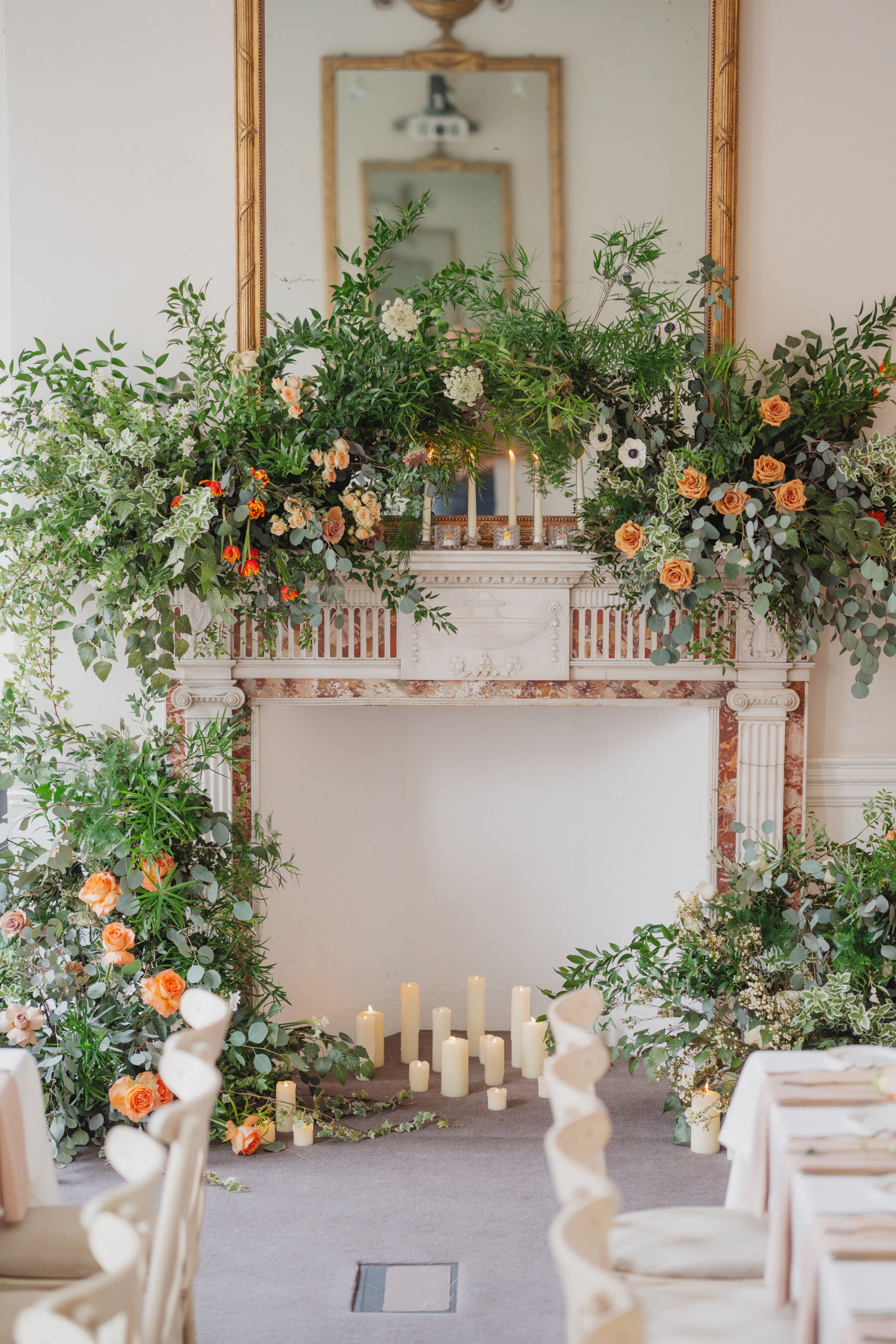 Sprawling foliage and orange flowers decorating the fireplace and mantlepiece at 41 Portland Place London. Candles of various height sit inside the fireplace. 