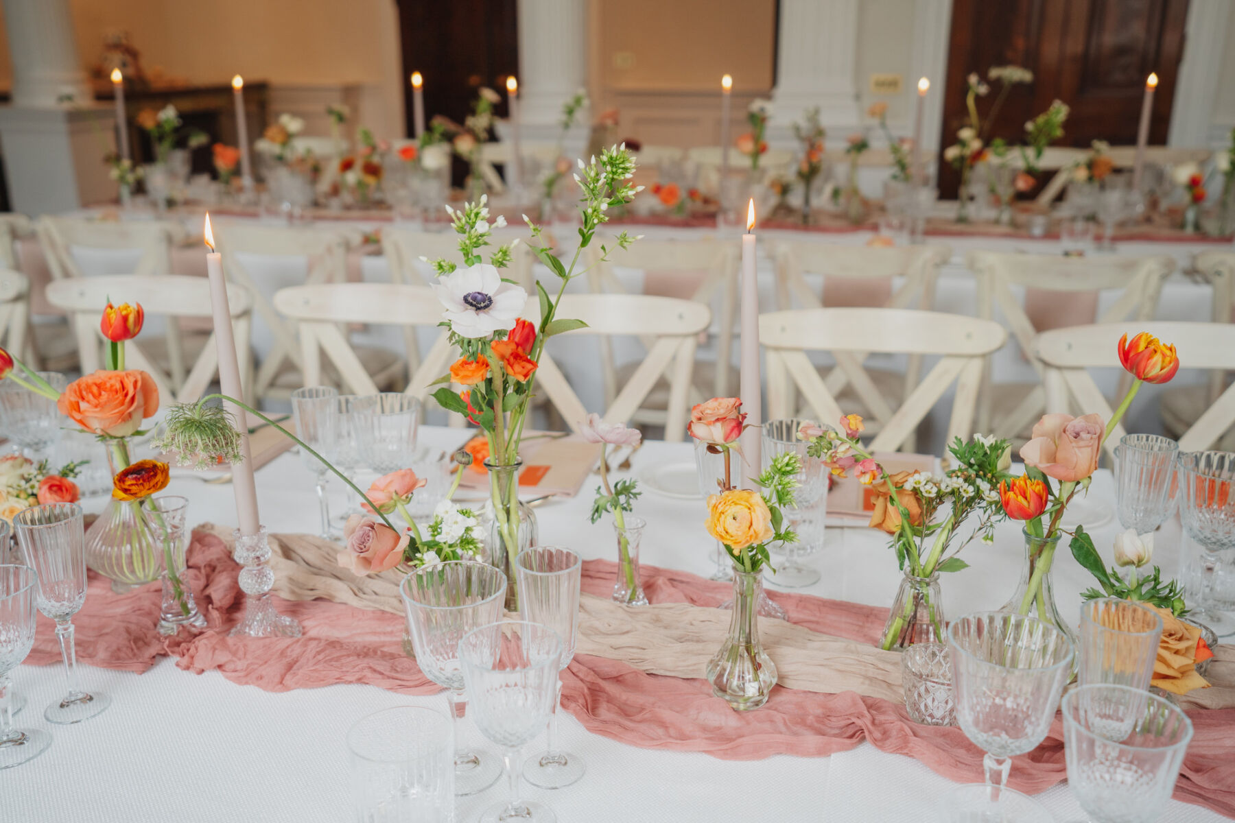 Elegant orange and cream wedding flowers in bud vases surrounded by rubbed glasses, taper candles and hand dyed table runners.