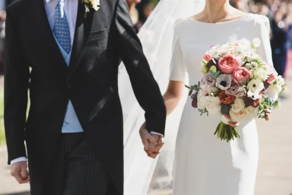 A cropped image of a bride and groom holding hands. The brides bouquet is visible and full of spring flowers.