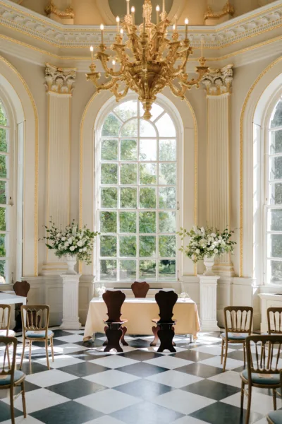 The image shows a wedding ceremony set up at Orleans House Gallery. A large orange window fills the image with two urns full of white flowers framing a table and chairs that sit just in front and below the large window.